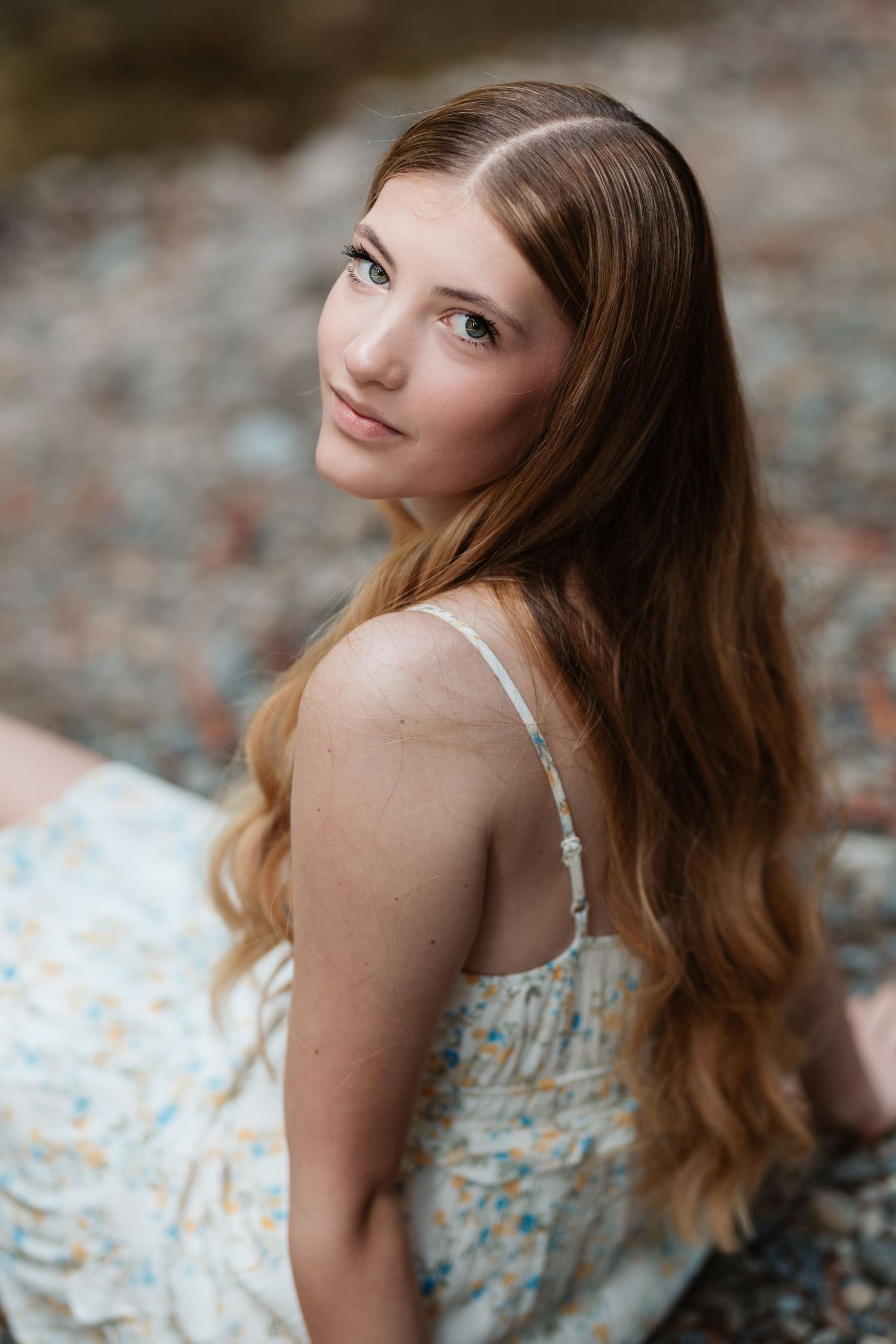 Young woman with long auburn wavy hair in a floral sundress sitting outdoors by rocky terrain.