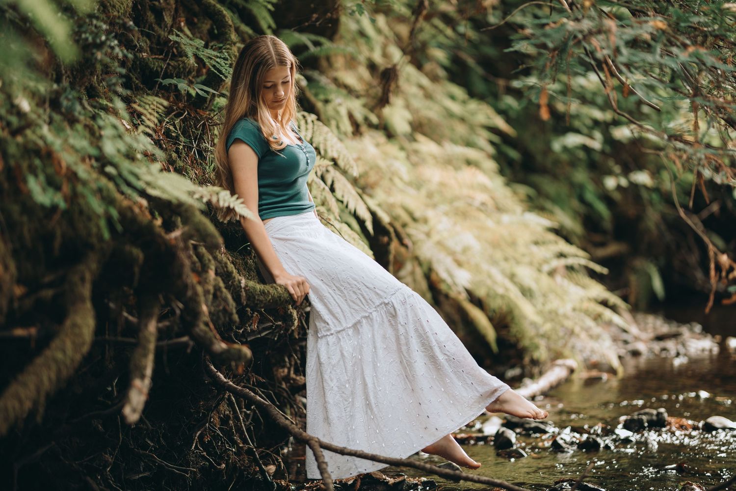 Young woman in teal top and white maxi skirt resting on tree roots along a forest creek.