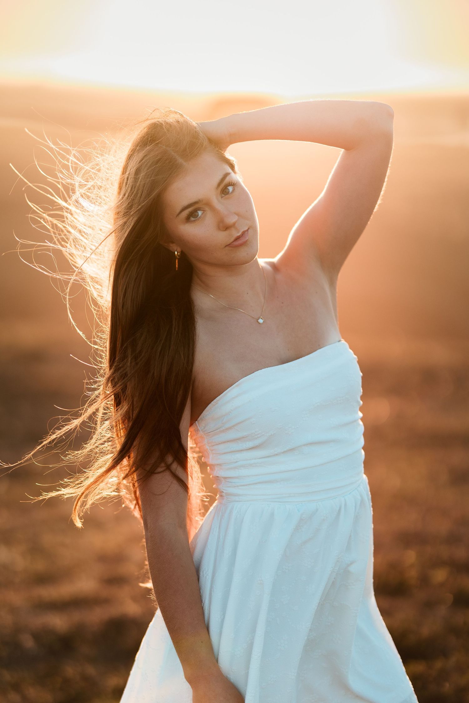Young woman in a white strapless dress posing in a field at sunset with wind blowing her long dark hair.