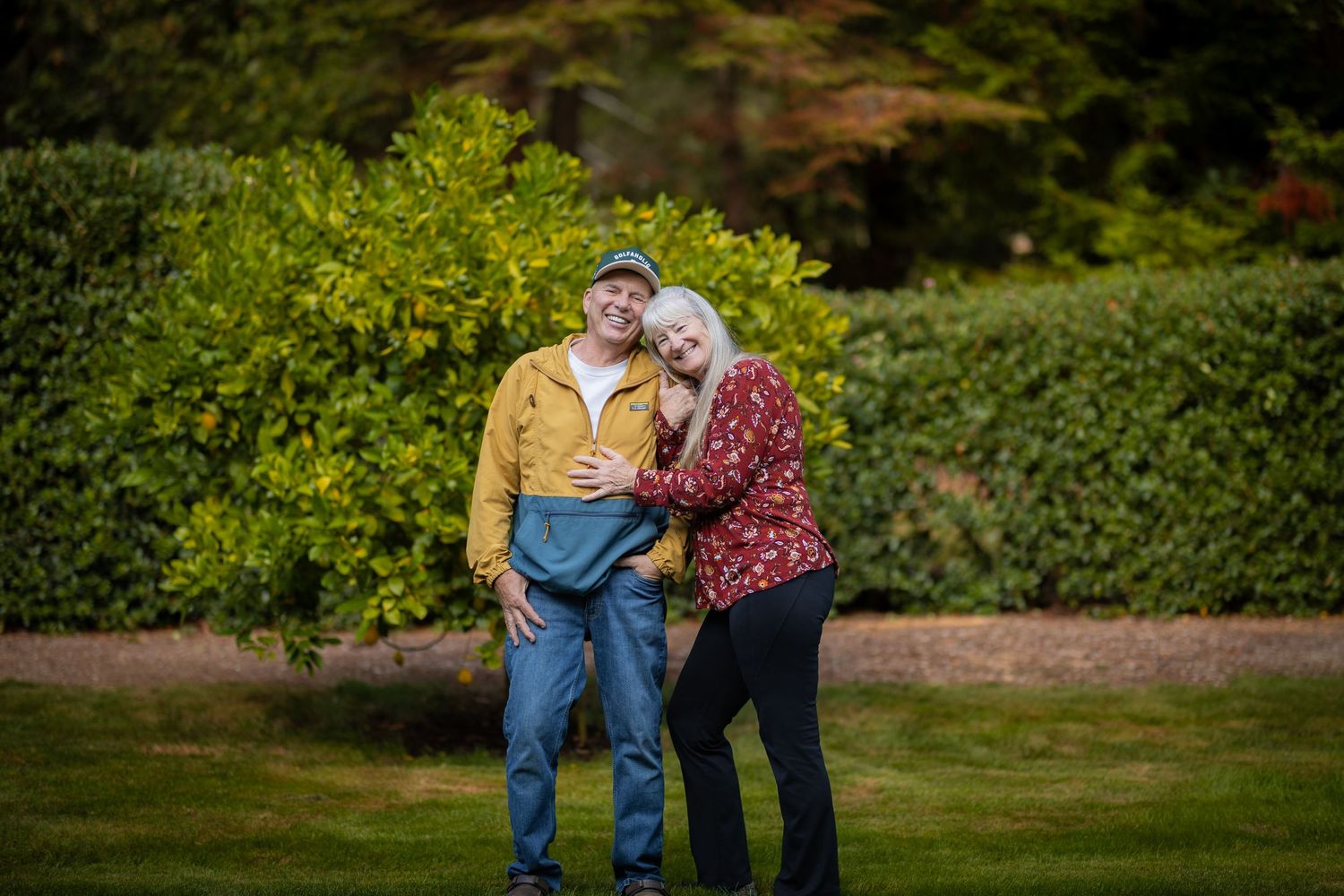 Smiling older couple embracing outdoors in a lush green garden, man in yellow jacket and woman in red floral top.