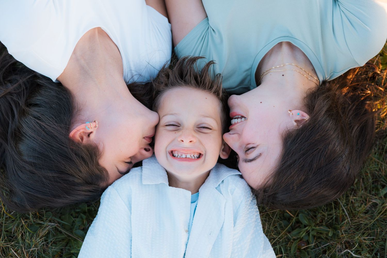 Two siblings kissing their smiling little brother on the cheeks while lying together on green grass outdoors.
