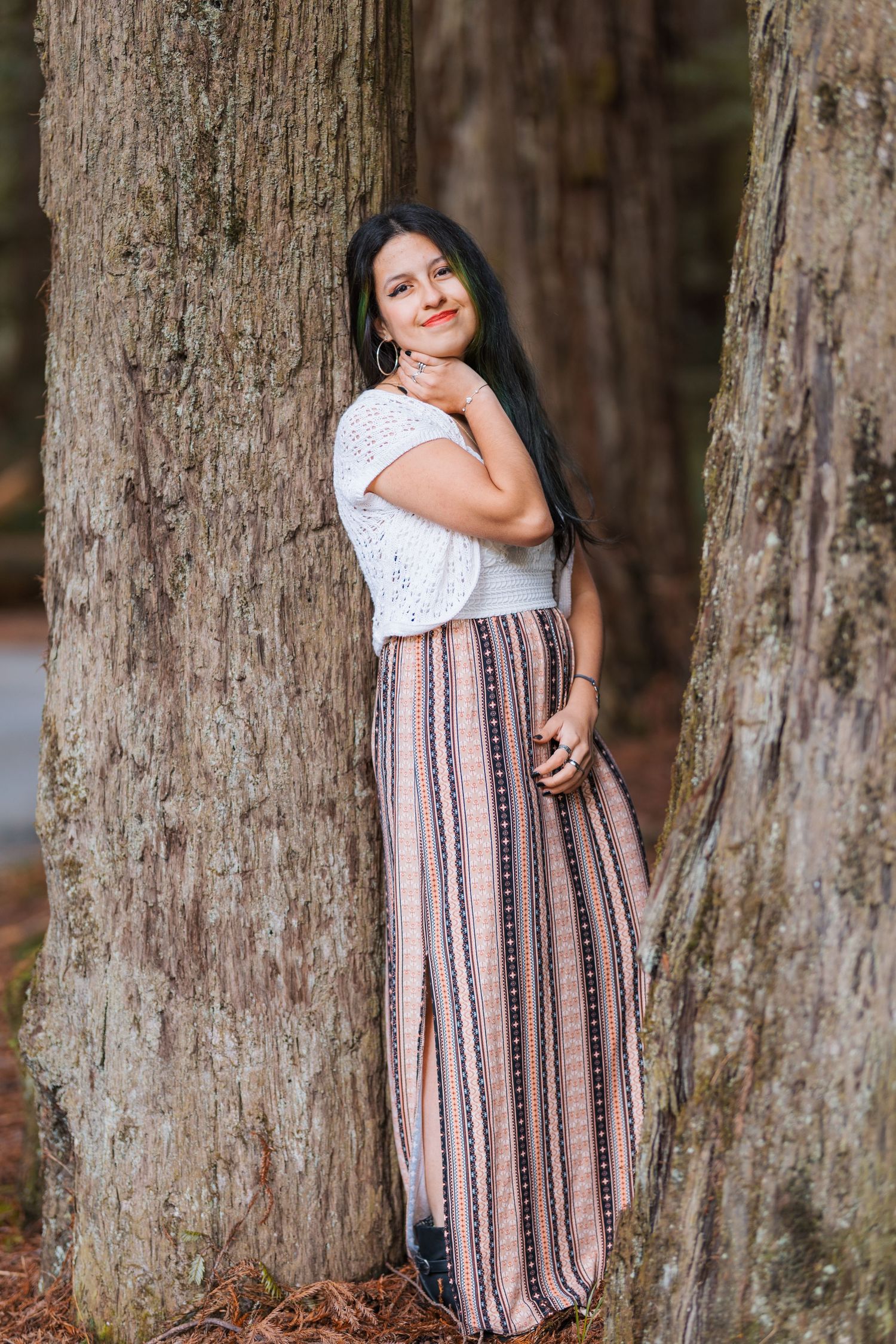 Young woman in white lace top and striped maxi skirt leans between two large tree trunks smiling.