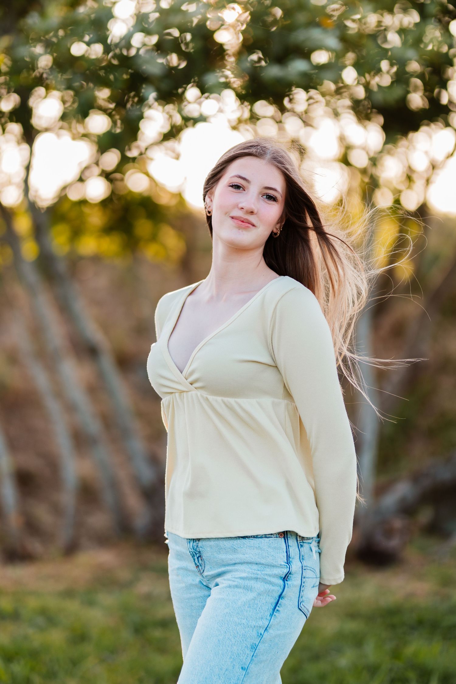 Young woman in yellow top and jeans smiles outdoors at golden hour with trees in background.