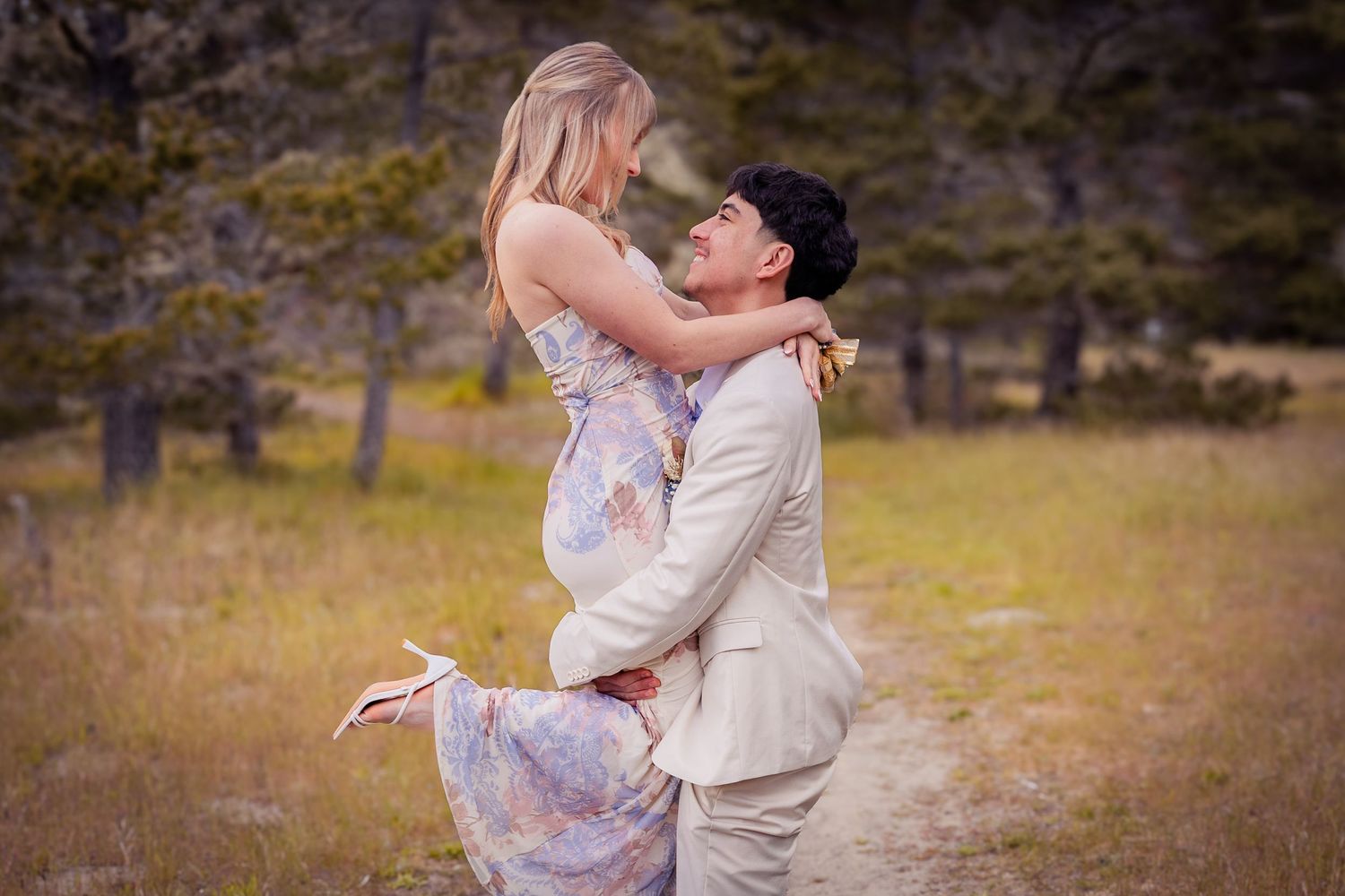 Couple embracing in a golden meadow, man lifting woman in floral dress during romantic outdoor engagement photo session.