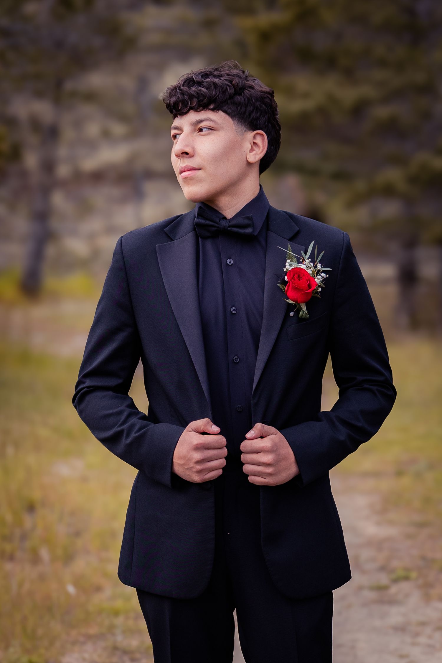 Young man in black tuxedo with red rose boutonniere and bow tie poses outdoors for formal occasion.
