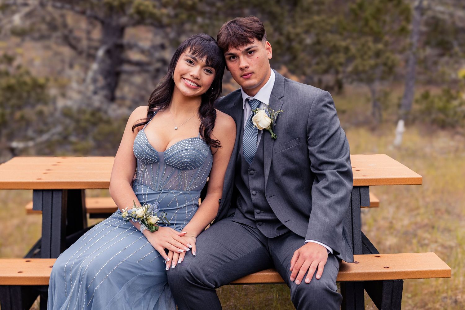 A young couple dressed in prom attire sit together on a wooden picnic bench outdoors, smiling for a photo.