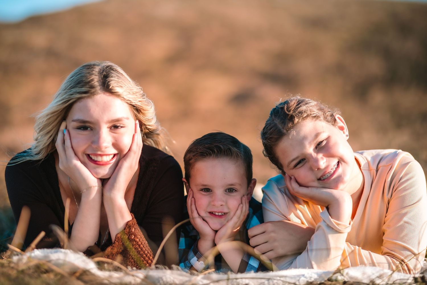 Three smiling children lying on the ground outdoors, posing with hands on cheeks in warm golden sunlight.
