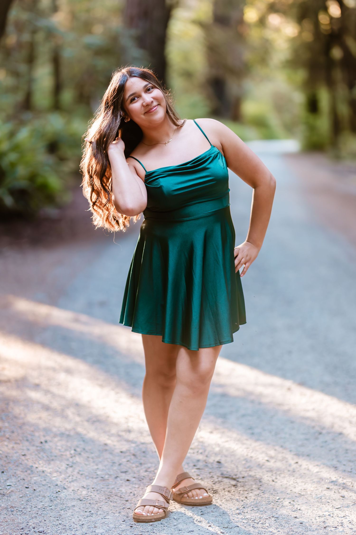 Young woman in green satin mini dress and sandals posing on a forest path, smiling confidently.