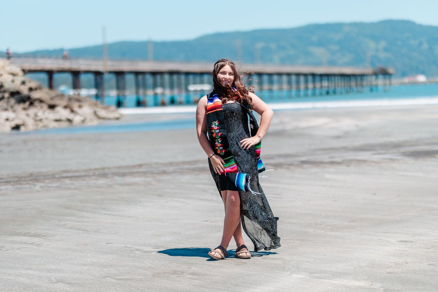 Woman in black dress holding sheer scarf poses on beach near pier on sunny day.