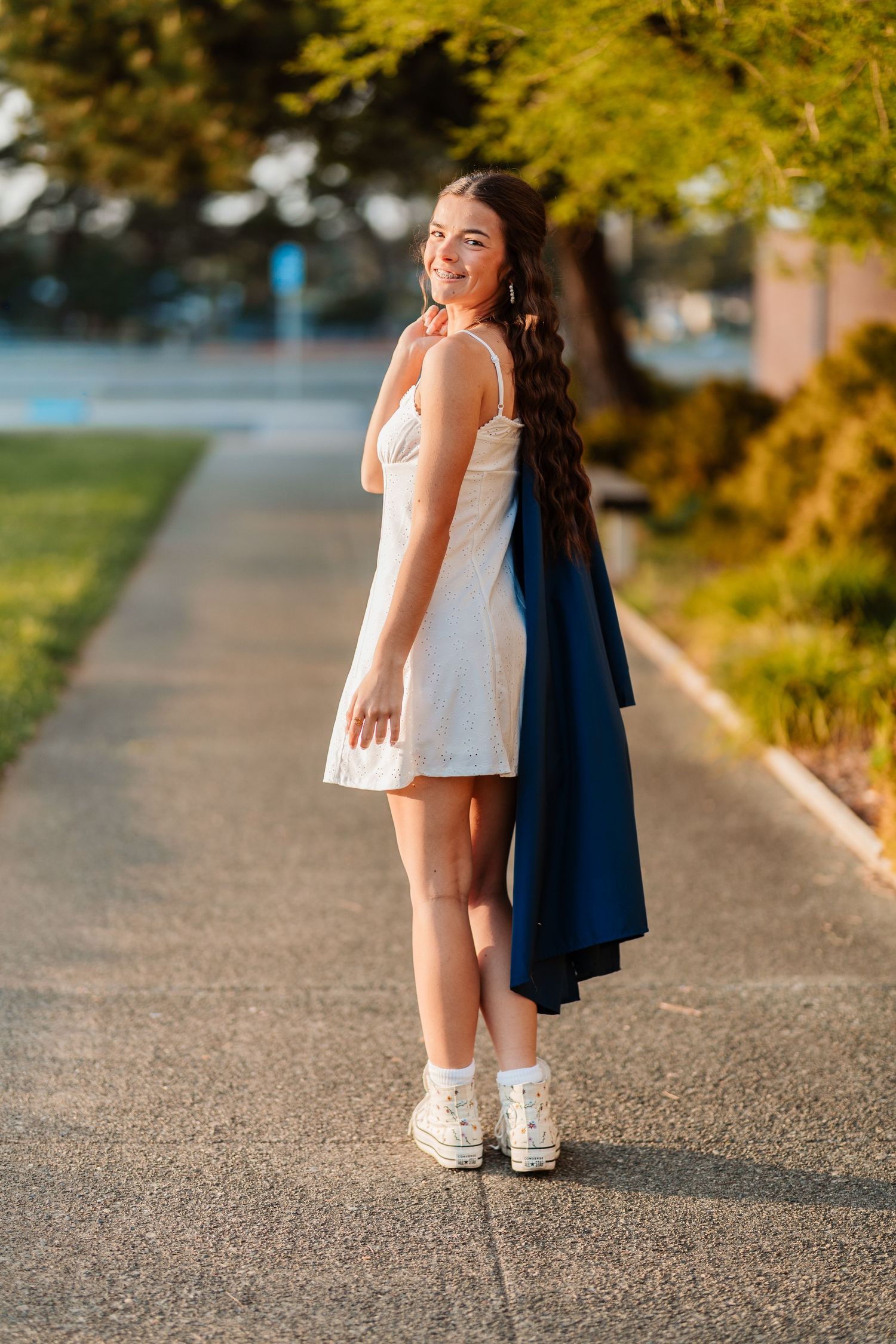 Smiling young woman in white dress and sneakers holding blue jacket on sunny outdoor pathway.