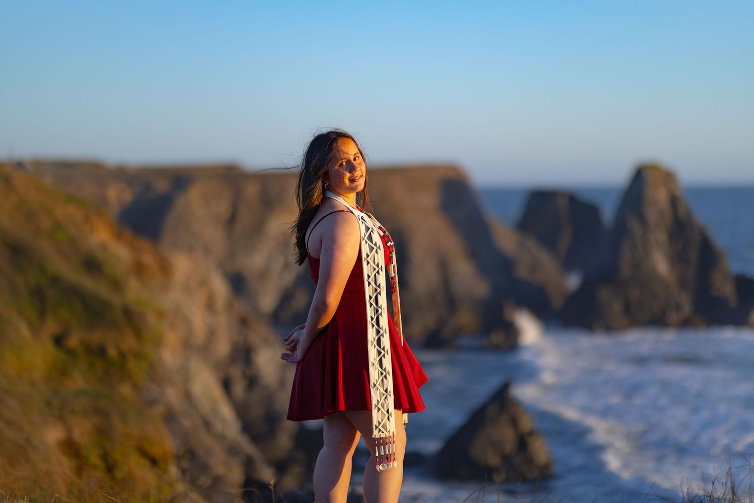 Young woman in red dress smiling on rocky coastal cliffs at golden hour with ocean waves in background.