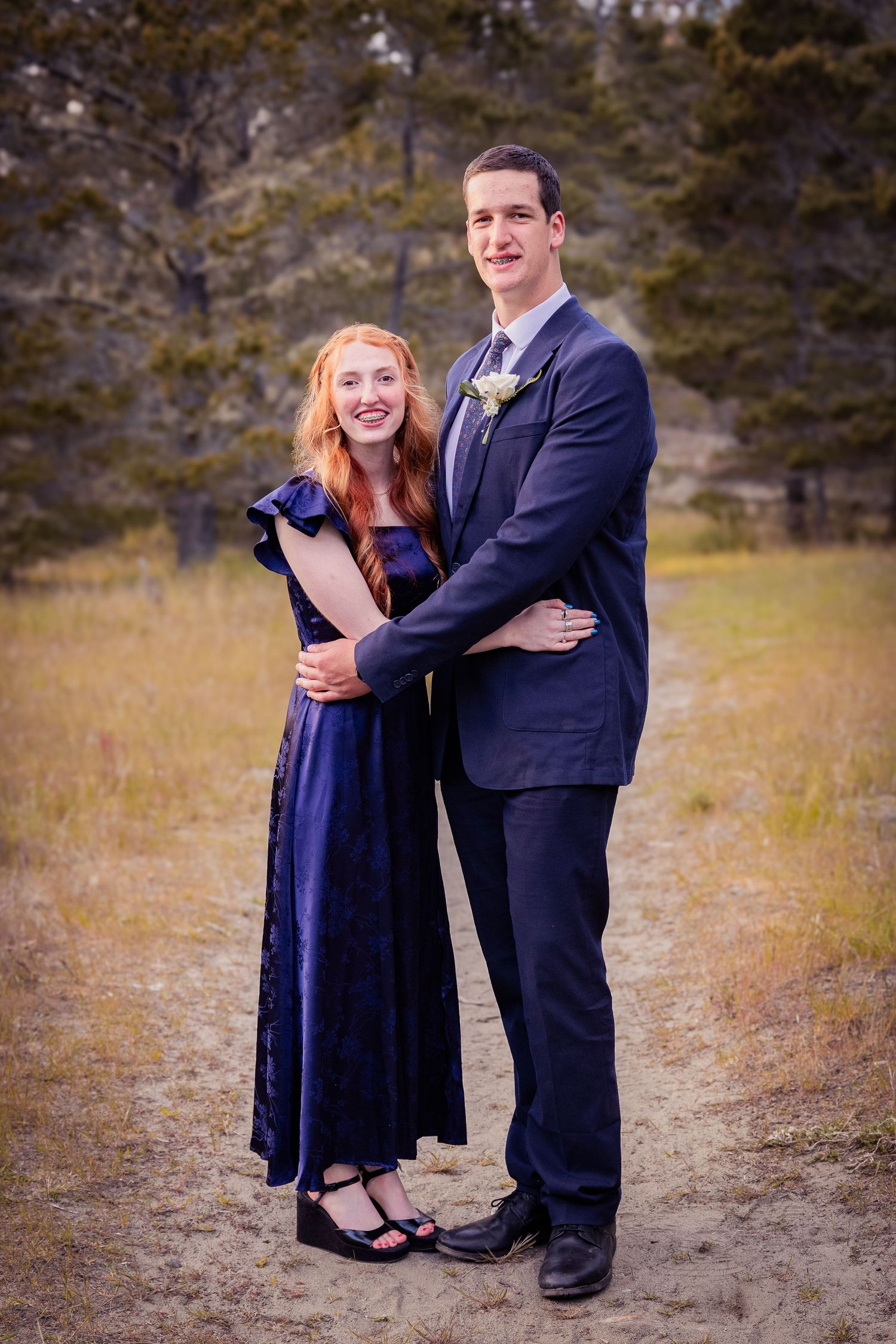 A smiling couple in formal prom attire pose together on a dirt path with trees in the background.