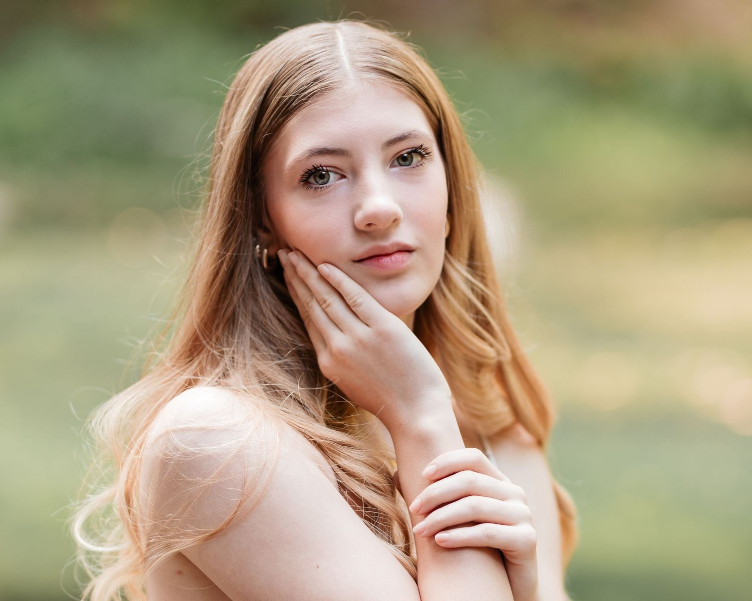 Close-up portrait of young woman with strawberry blonde hair touching face in soft outdoor lighting.