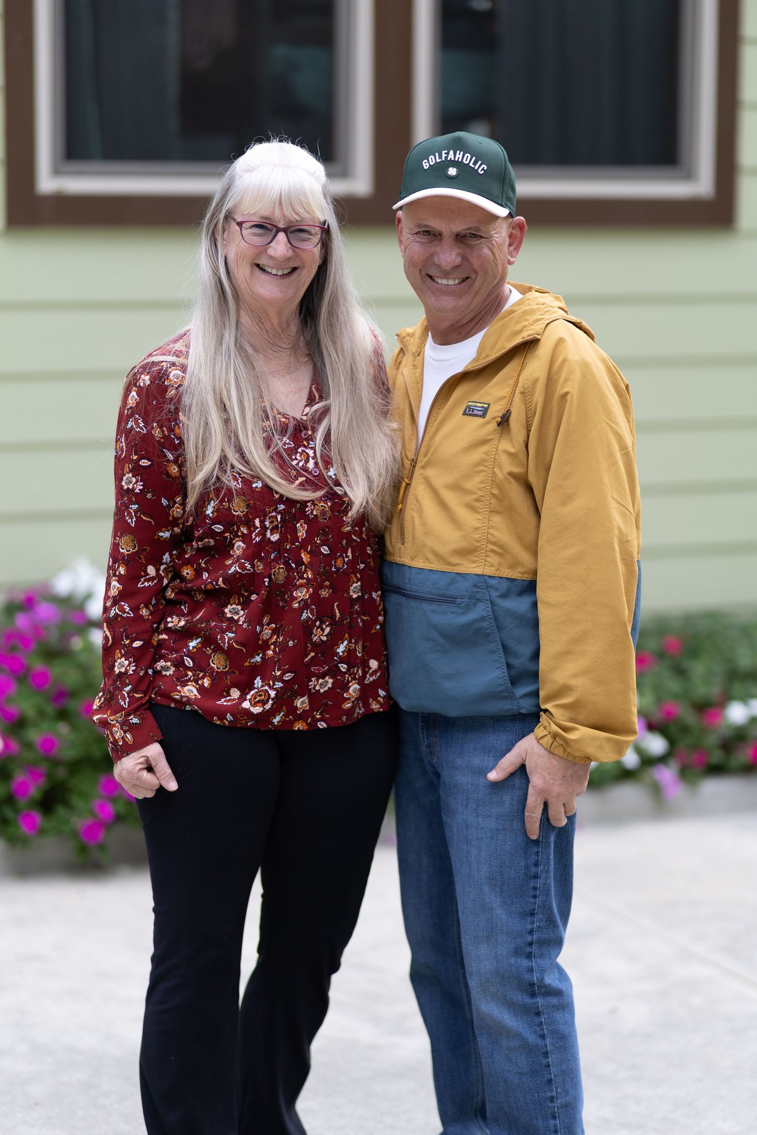 A smiling older couple standing together outdoors near a green building with flowers in background.