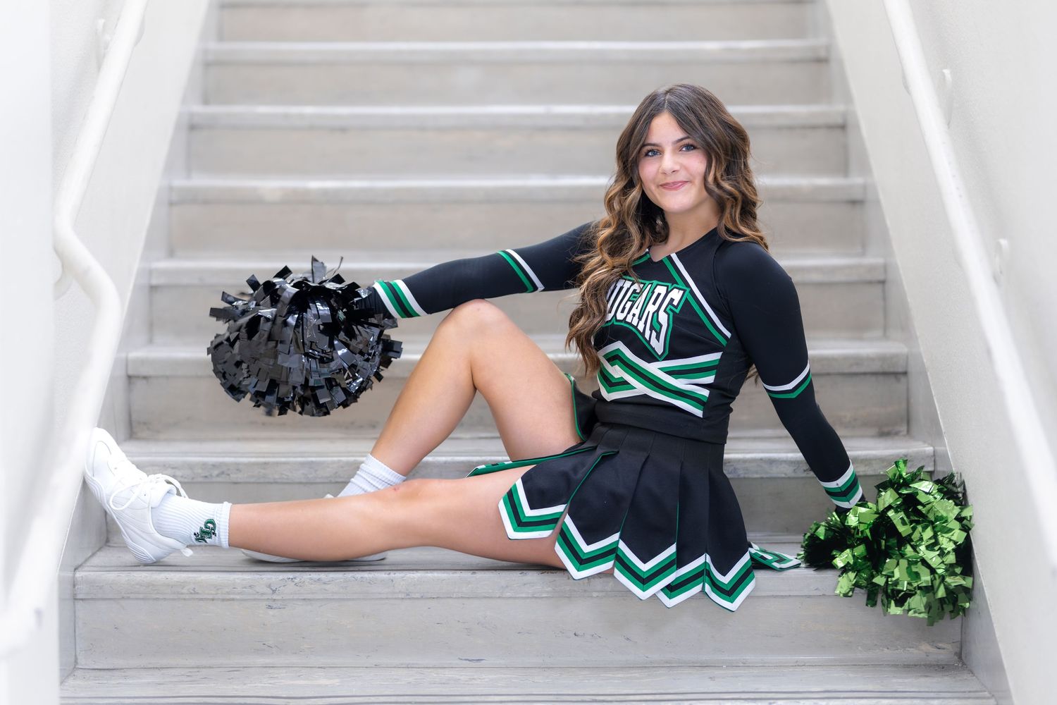 Cheerleader in black and green uniform sitting on stairs holding black and green pom-poms.