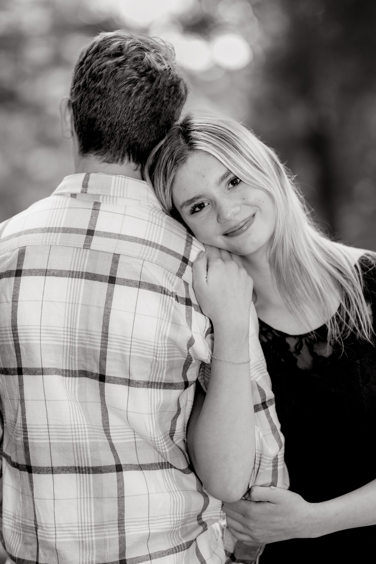 Black and white portrait of blonde woman resting head on mans shoulder embracing outdoors.