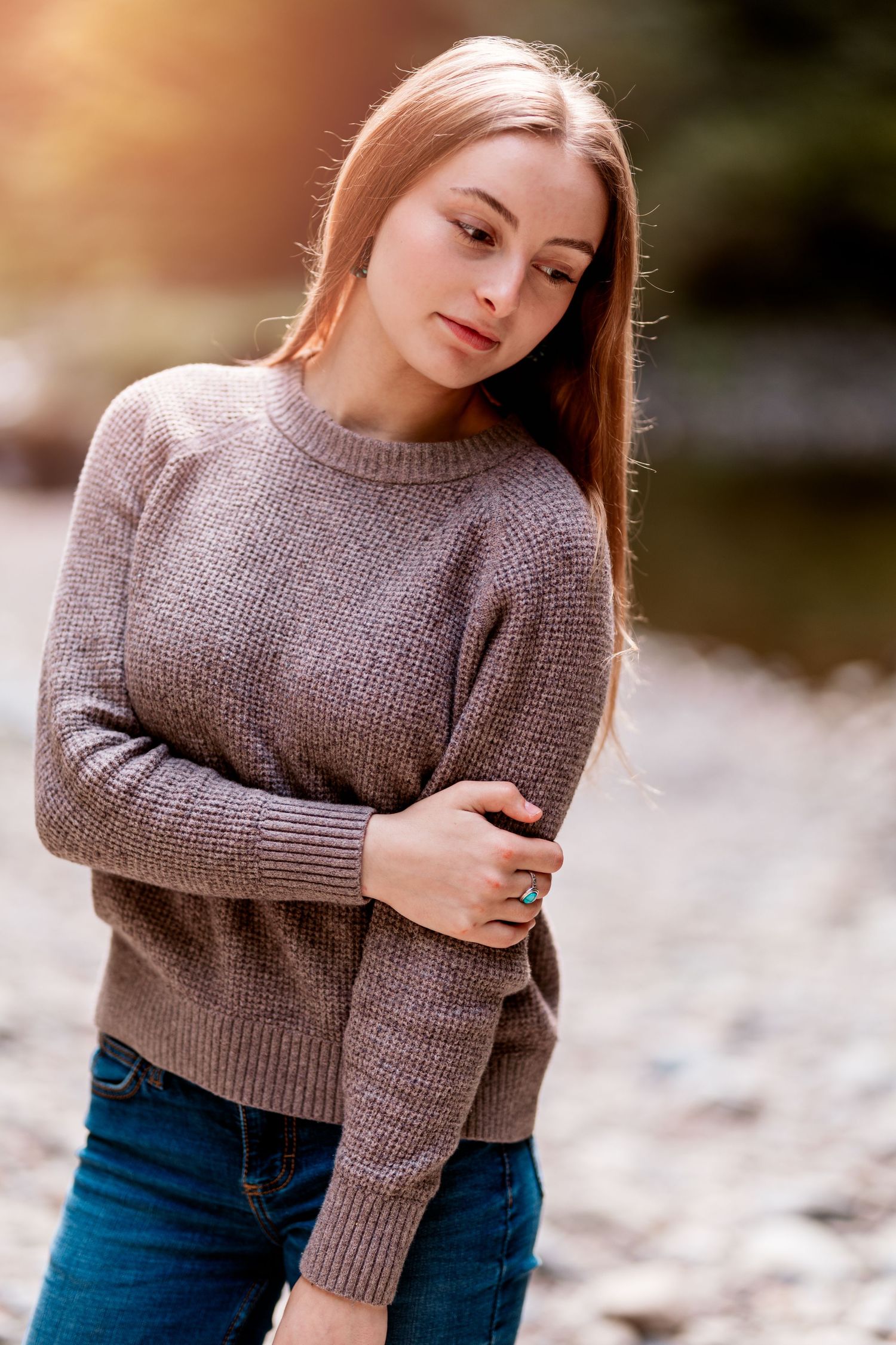 Young woman with long auburn hair in brown knit sweater and jeans posing outdoors by a rocky river.