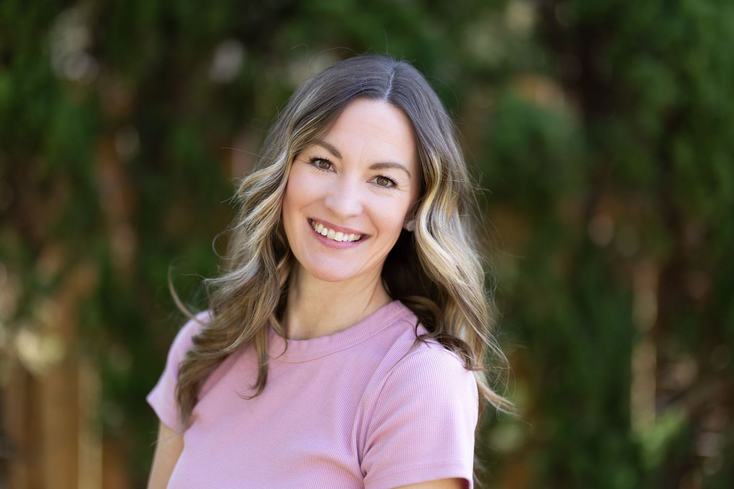 Smiling woman with wavy highlighted hair wearing a pink top, posing outdoors with green trees in the background.