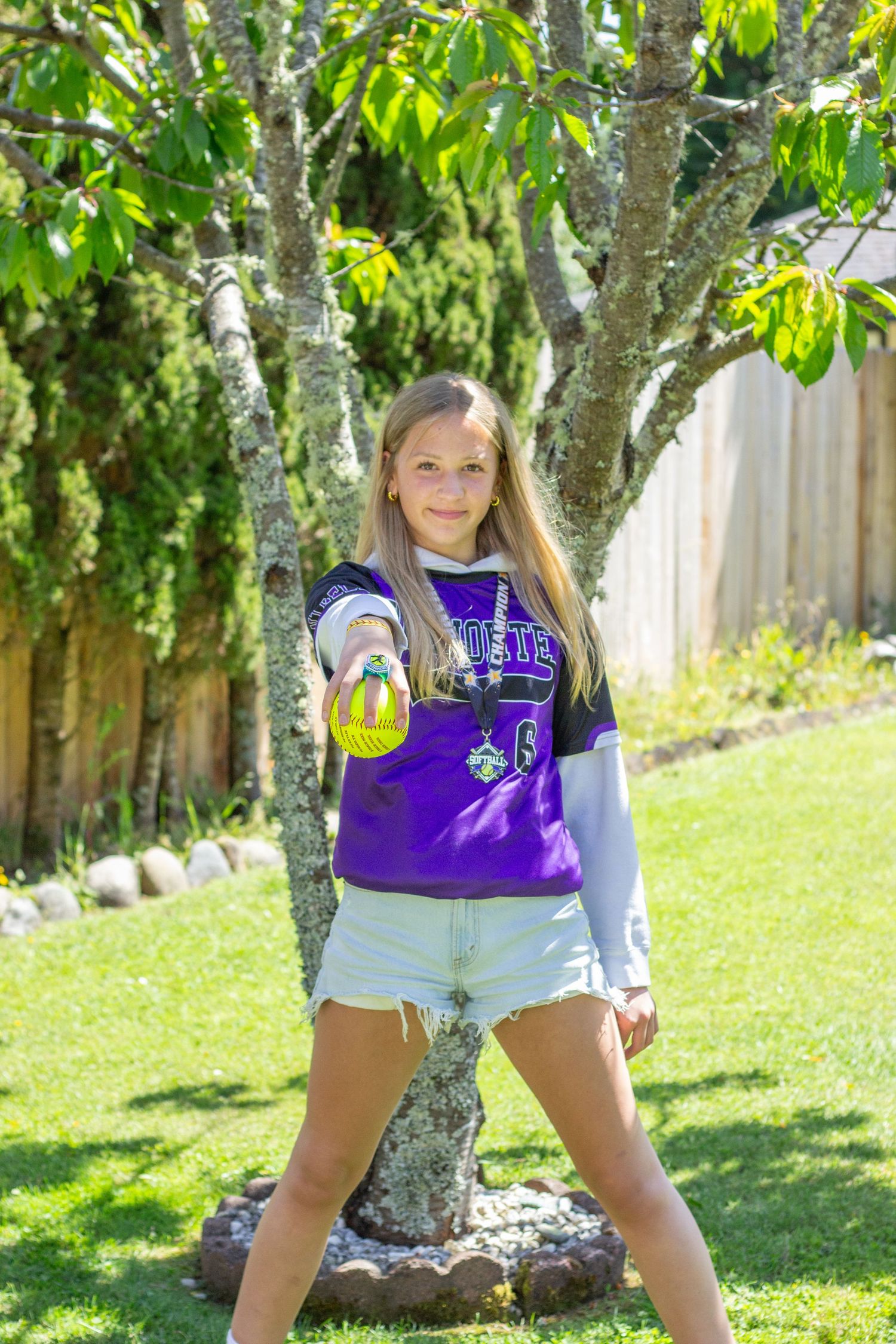 Young girl in purple softball jersey holding a yellow softball toward camera in sunny backyard.