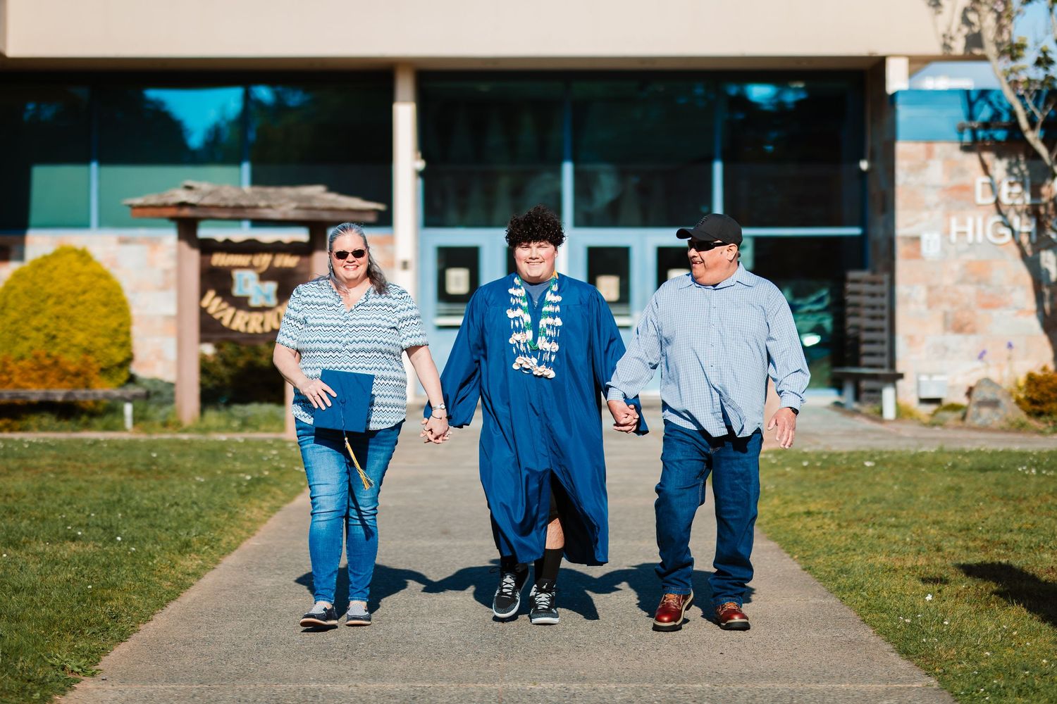 Graduate in blue gown walks hand-in-hand with proud parents outside school building on sunny day.