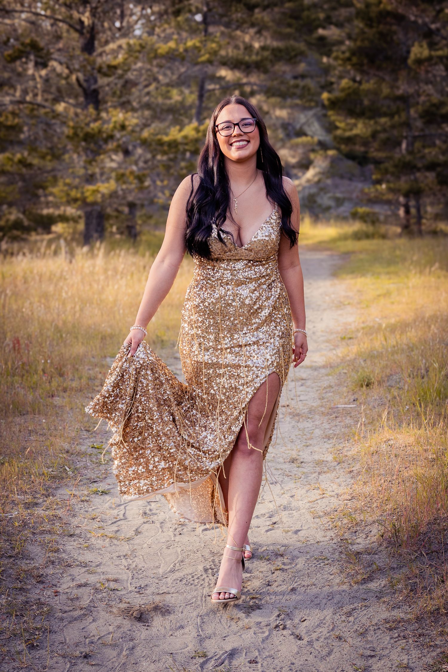 Young woman in gold sequin gown walking on sandy path through golden meadow, smiling joyfully.