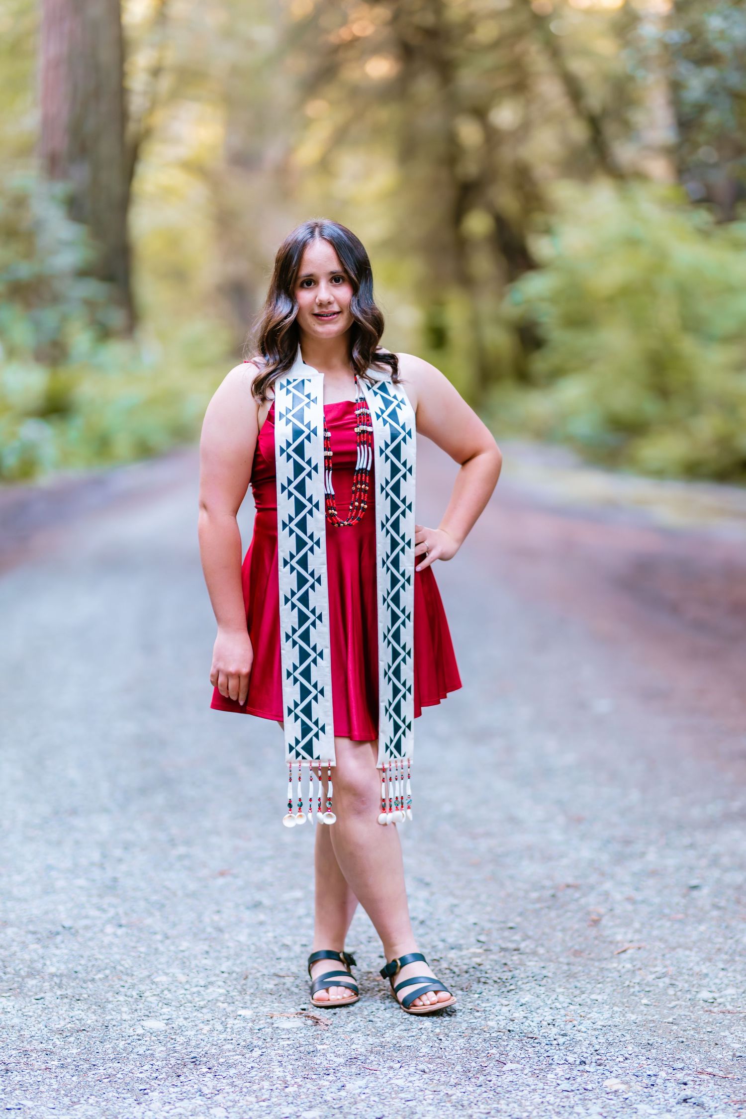 Young woman in red dress with decorative sash standing confidently on a tree-lined path in warm light.