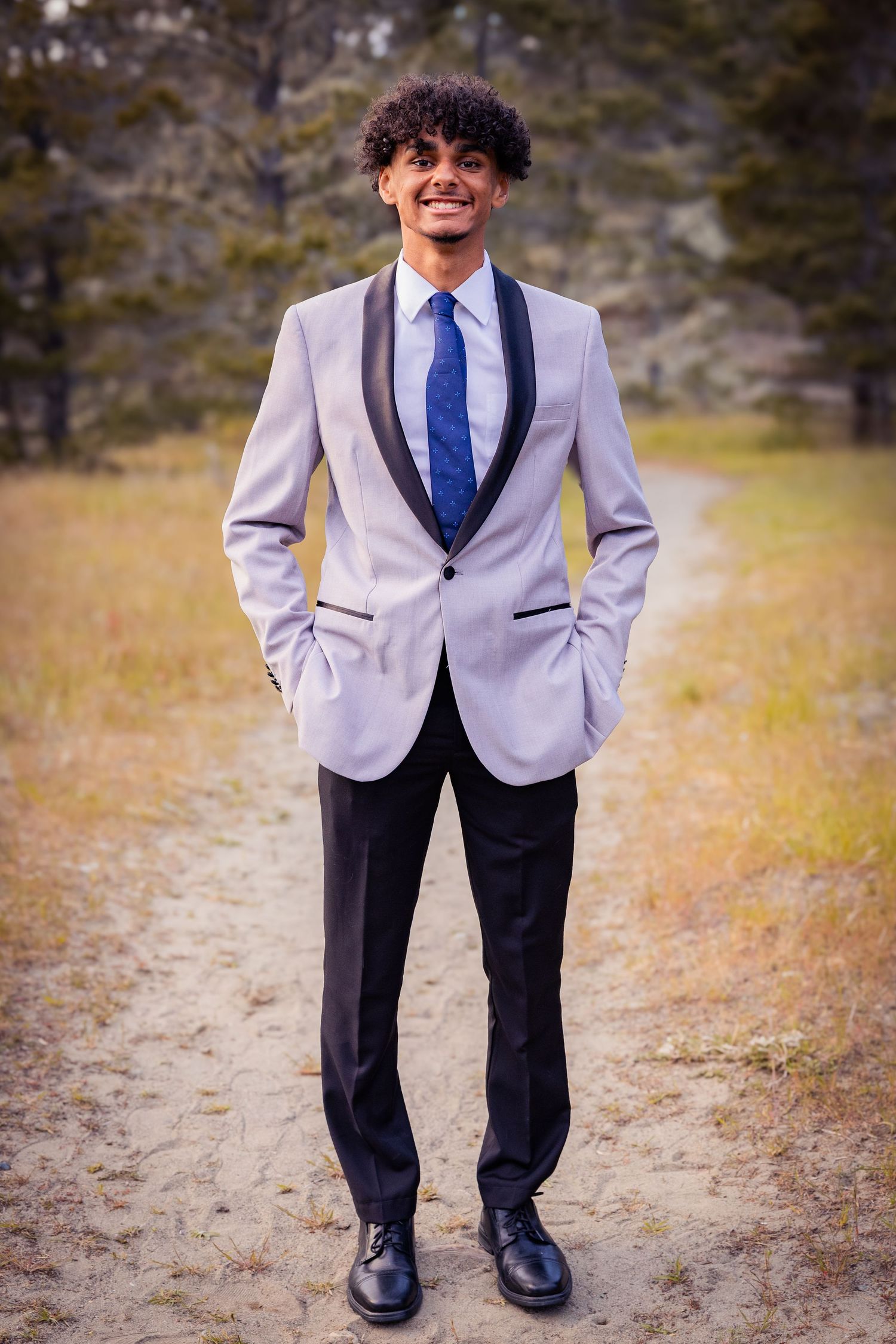 Young man in gray tuxedo jacket with black lapels, blue tie, and black pants smiling outdoors.