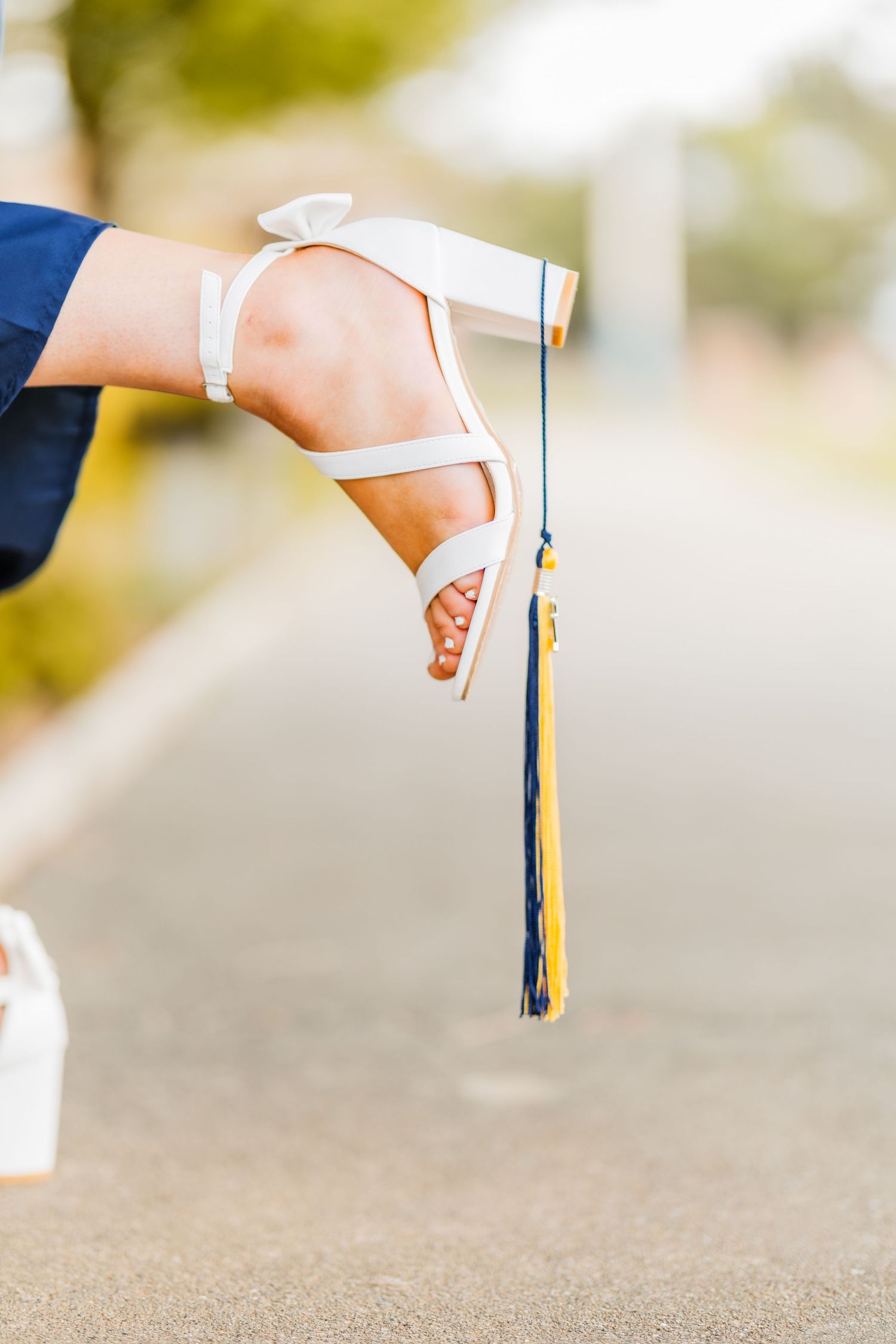Close-up of a woman's foot in a white heeled sandal holding a graduation tassel on a blurred outdoor background.