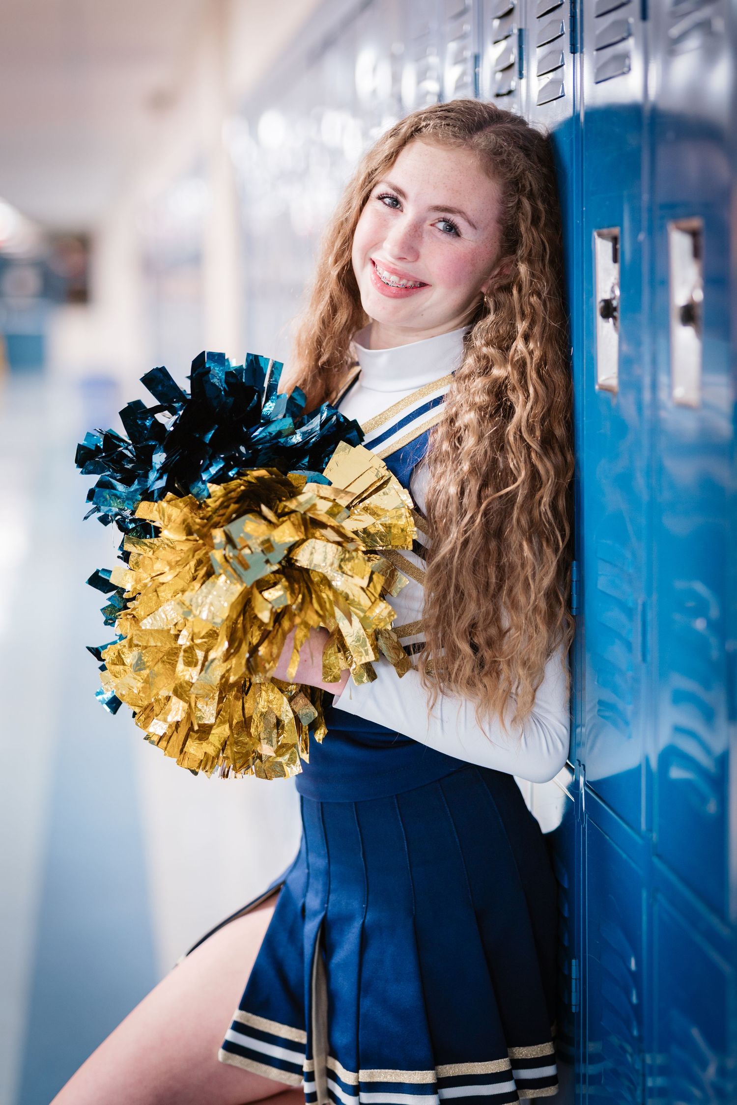 Cheerleader in blue and white uniform holding gold and teal pom-poms by school lockers.