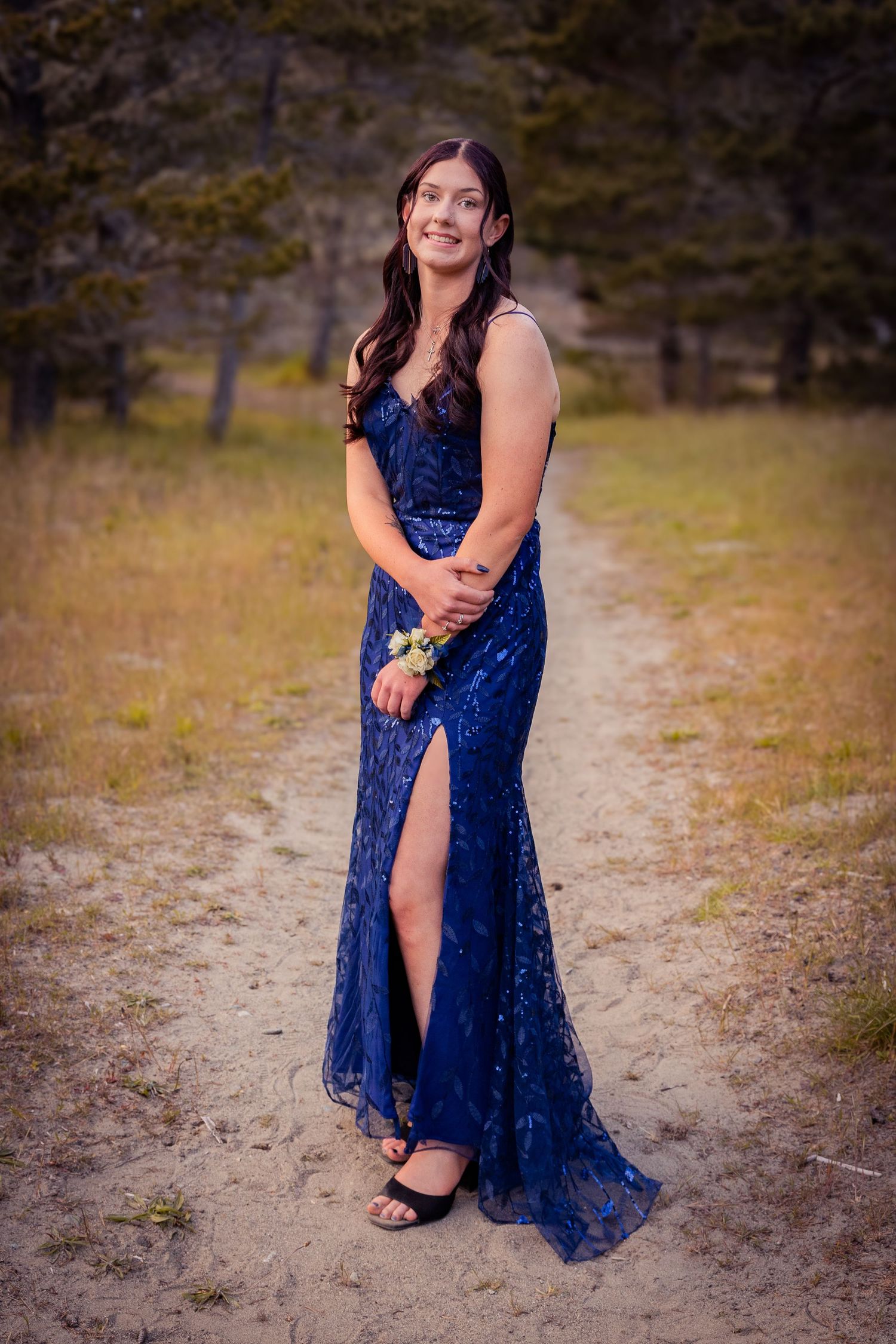 Young woman in a sequined navy blue prom dress with slit poses on a dirt path surrounded by trees.