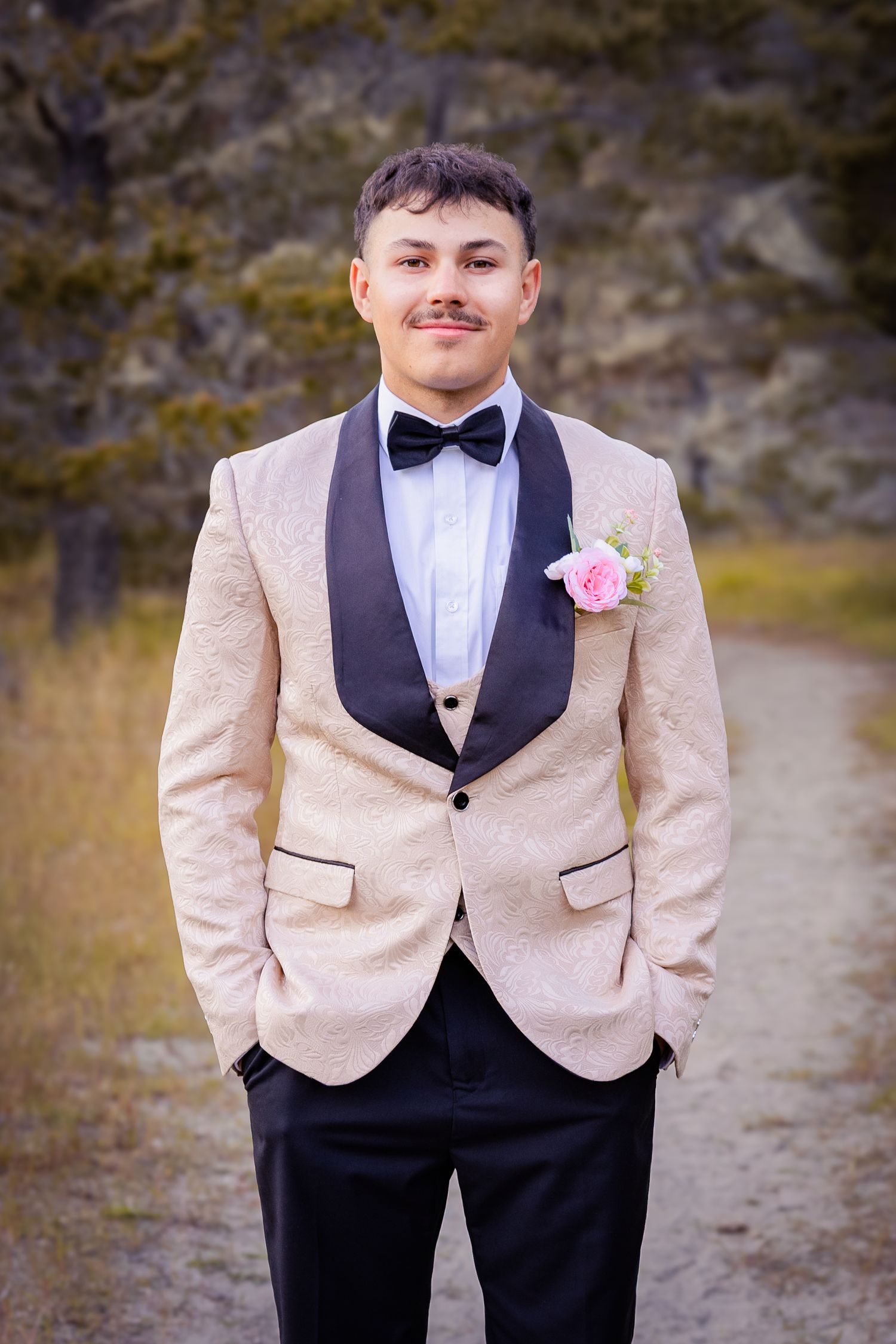 Young man in cream tuxedo jacket with black lapels, bow tie, and pink boutonniere poses outdoors on a gravel path.
