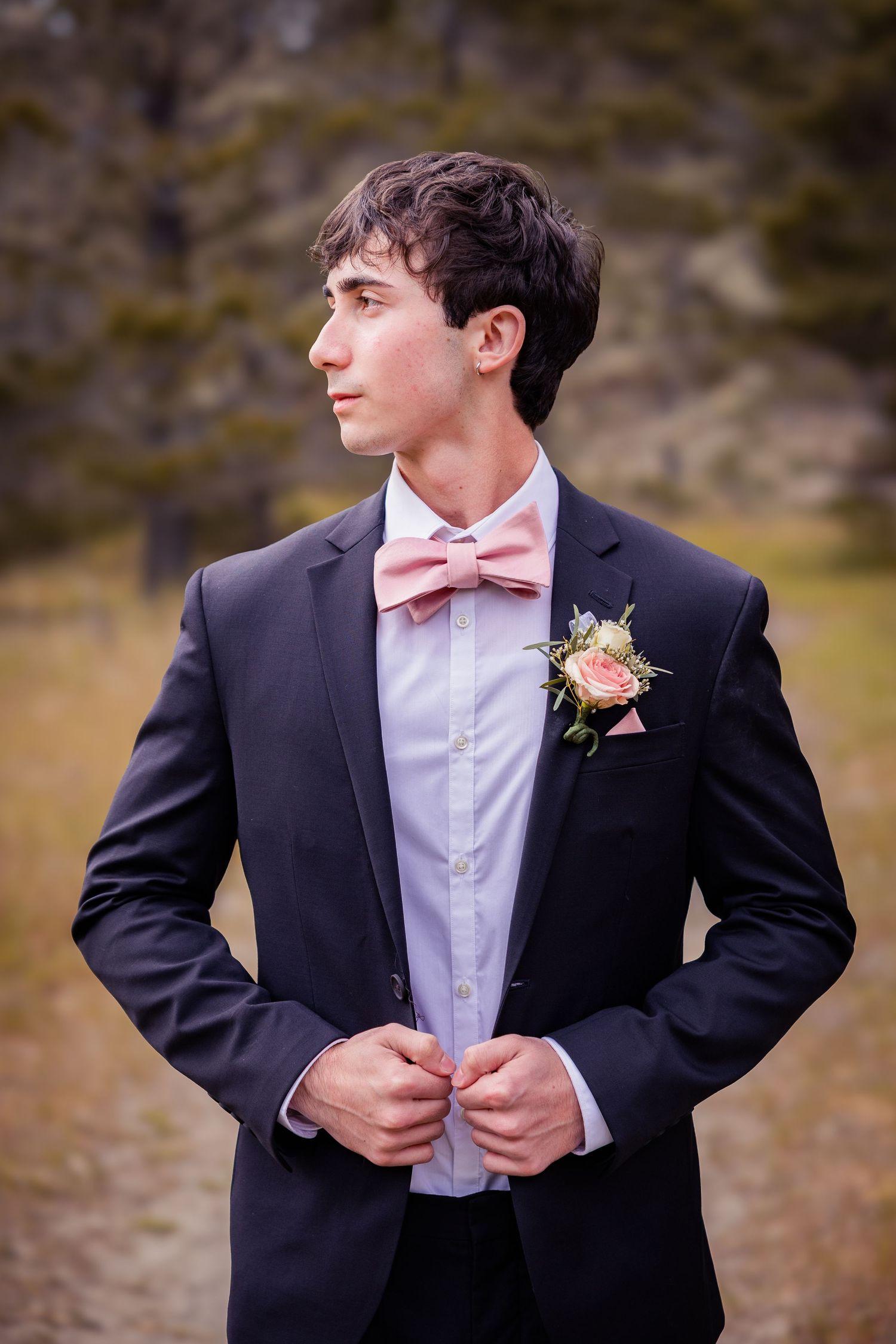 Young groom in black suit with pink bow tie and boutonniere buttoning jacket outdoors.