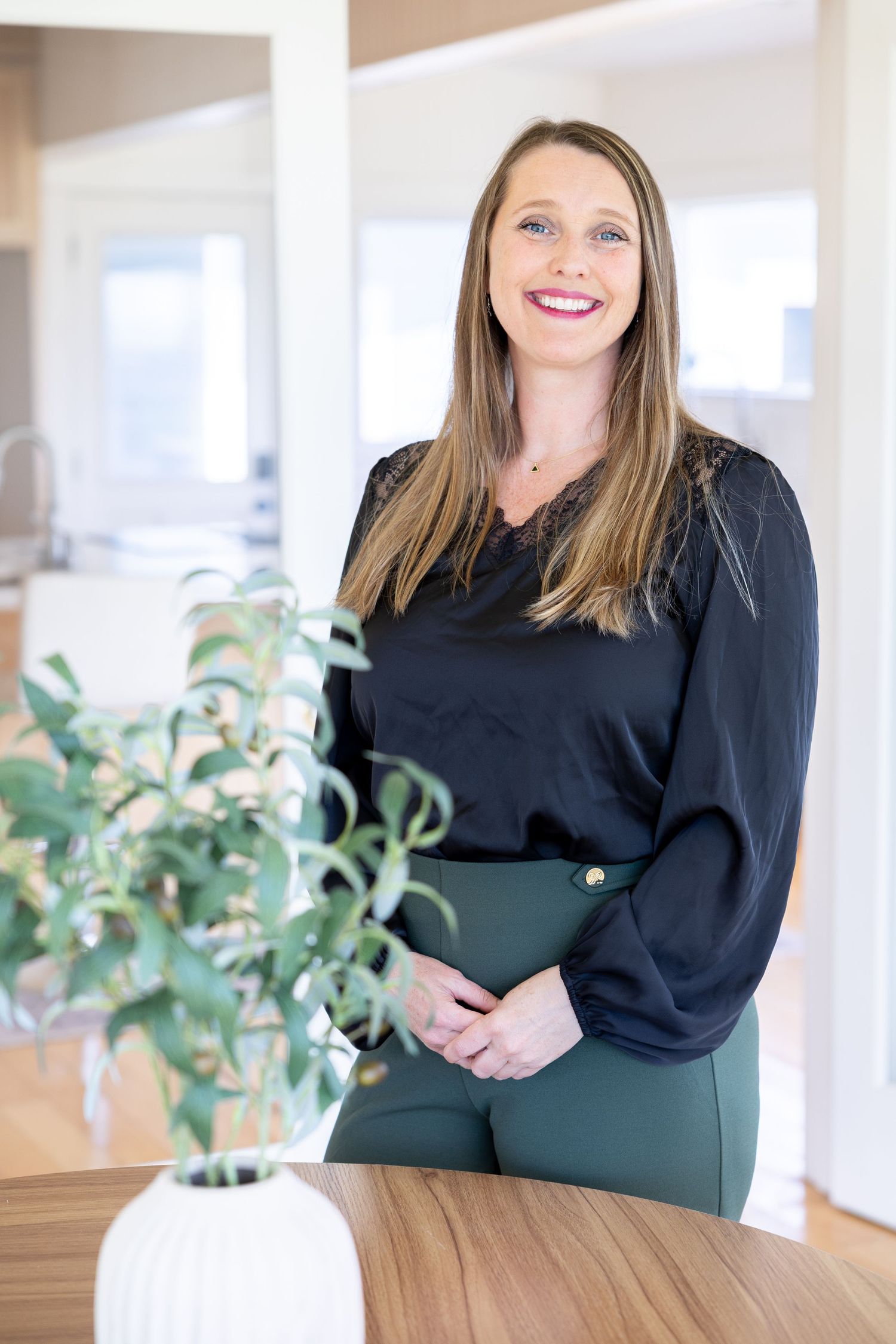 A smiling woman in a black blouse and green pants stands by a wooden table with a white vase in a modern home.