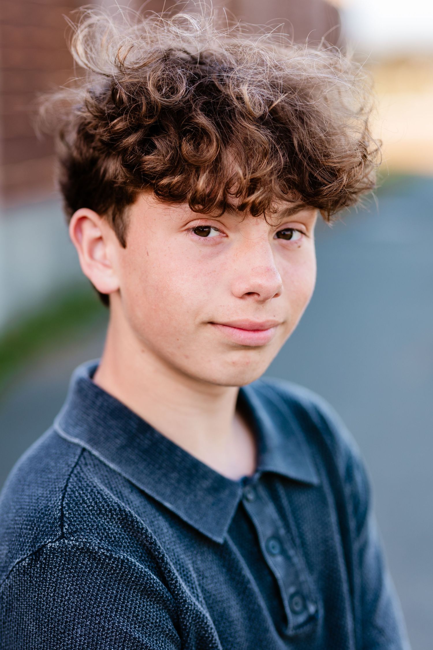 Teen boy with curly brown hair wearing a dark polo shirt, posing outdoors in a candid portrait photo.