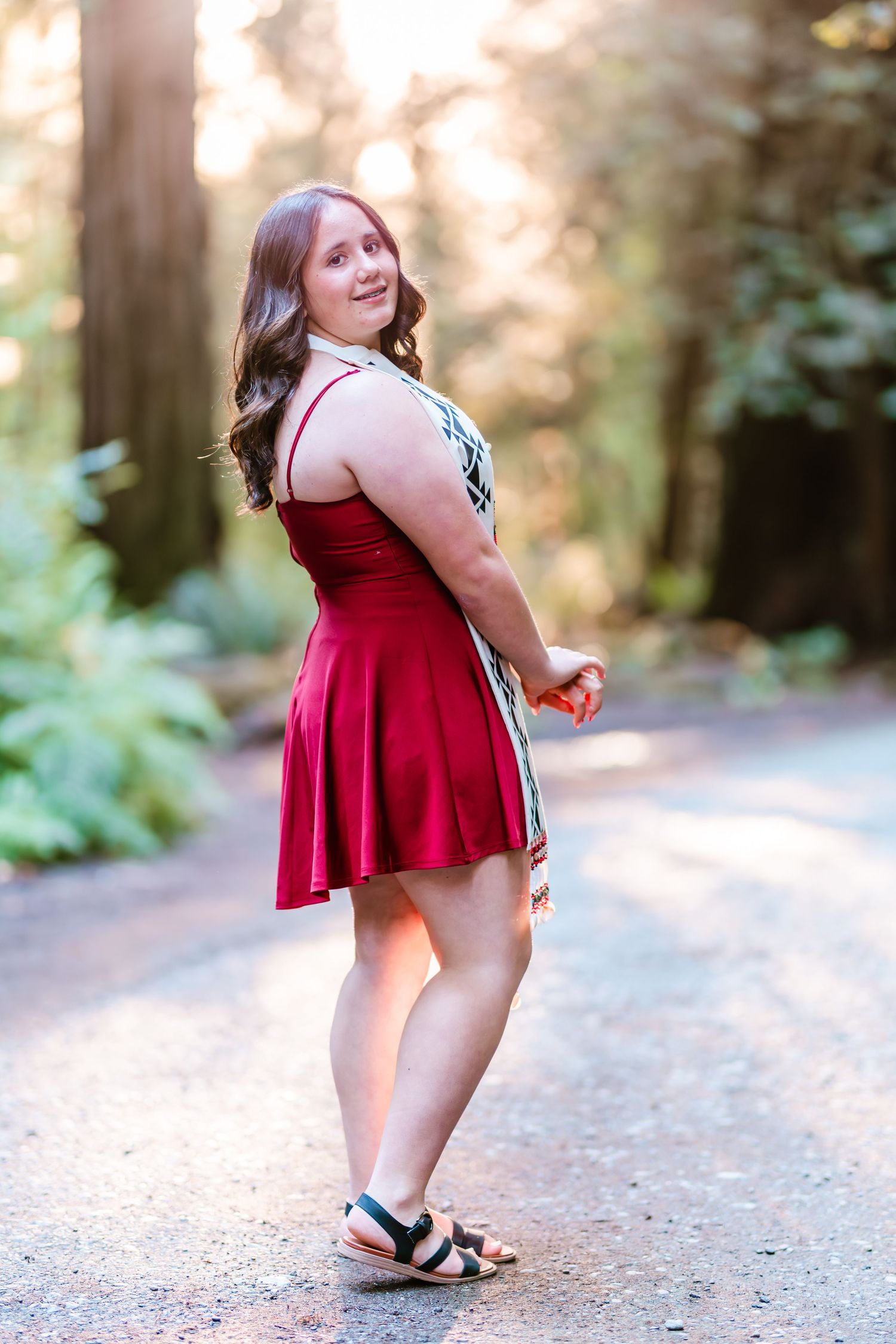 Young woman in red dress and sandals posing on a forest path with sunlight filtering through tall trees.