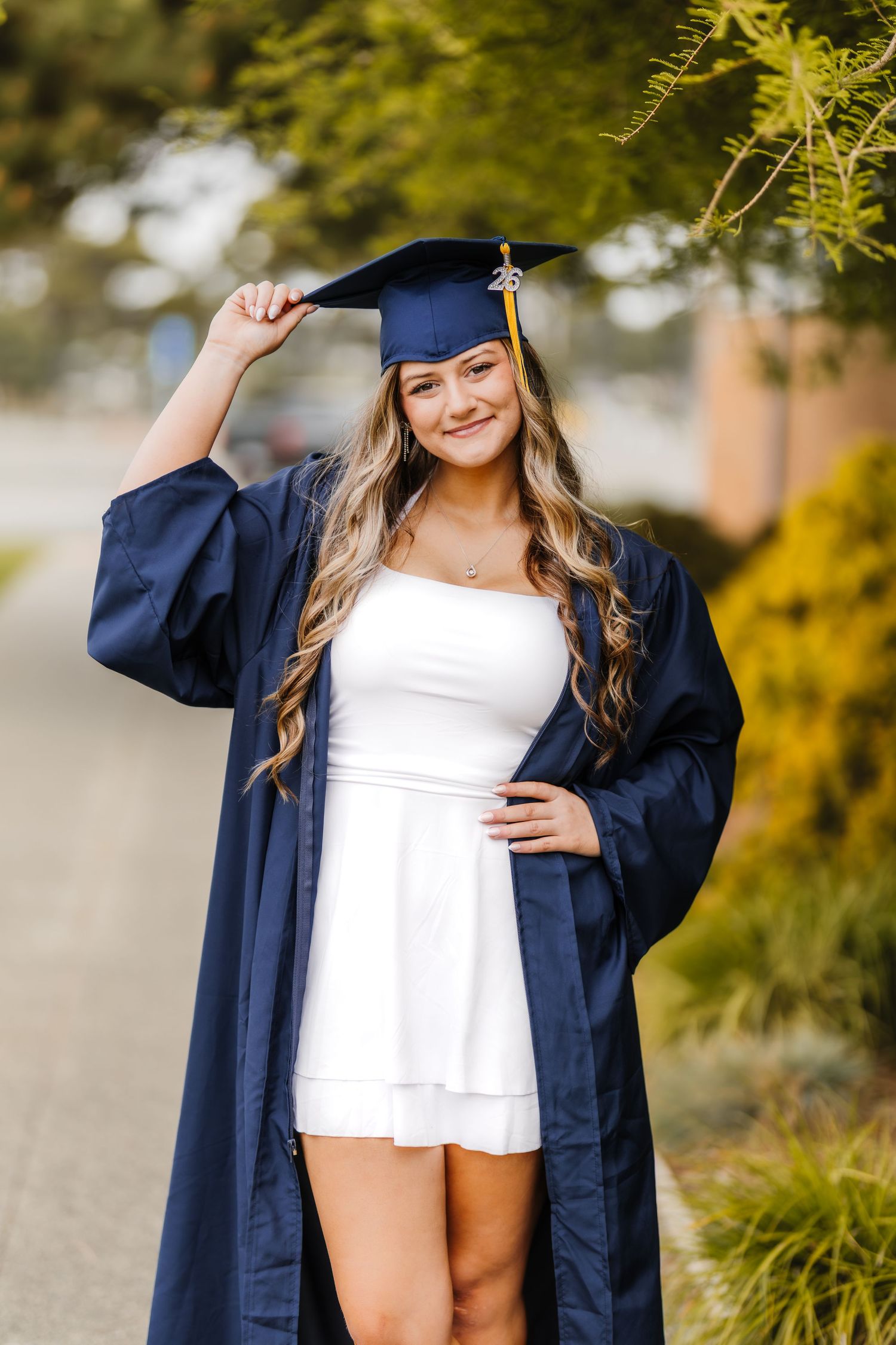 Smiling graduate in navy cap and gown over white dress, posing outdoors.
