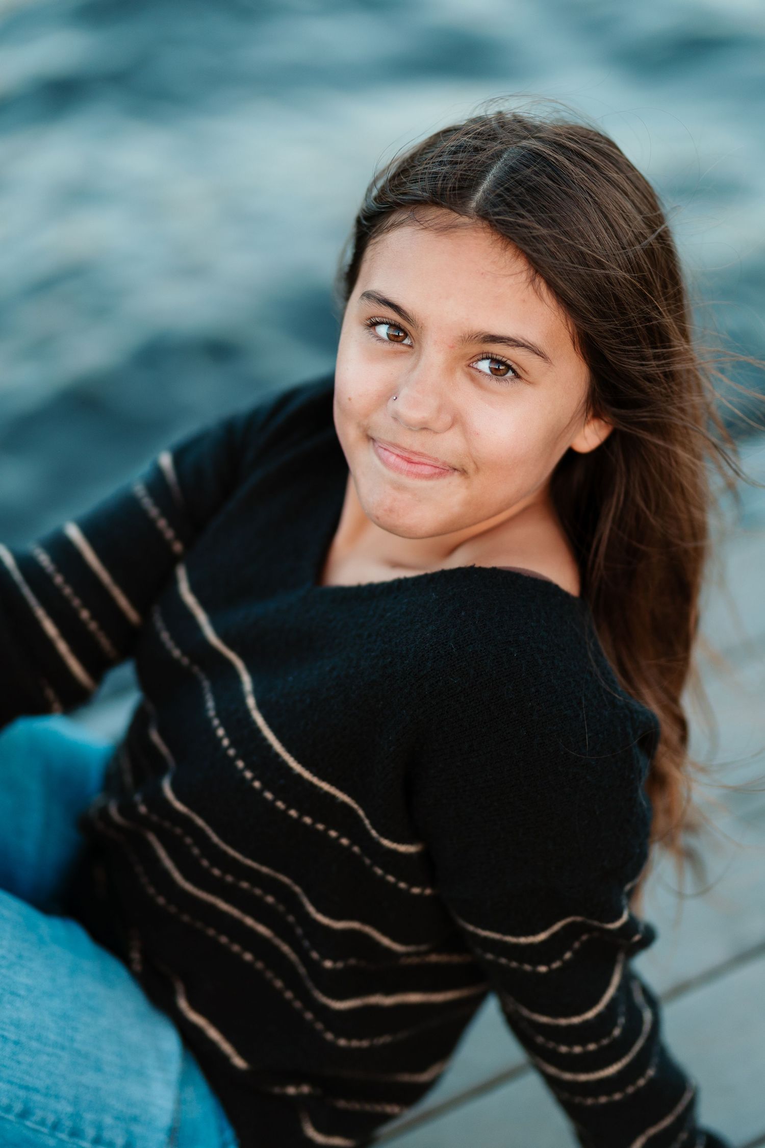 Young girl with dark hair smiling while seated near water in a black striped sweater at dusk.