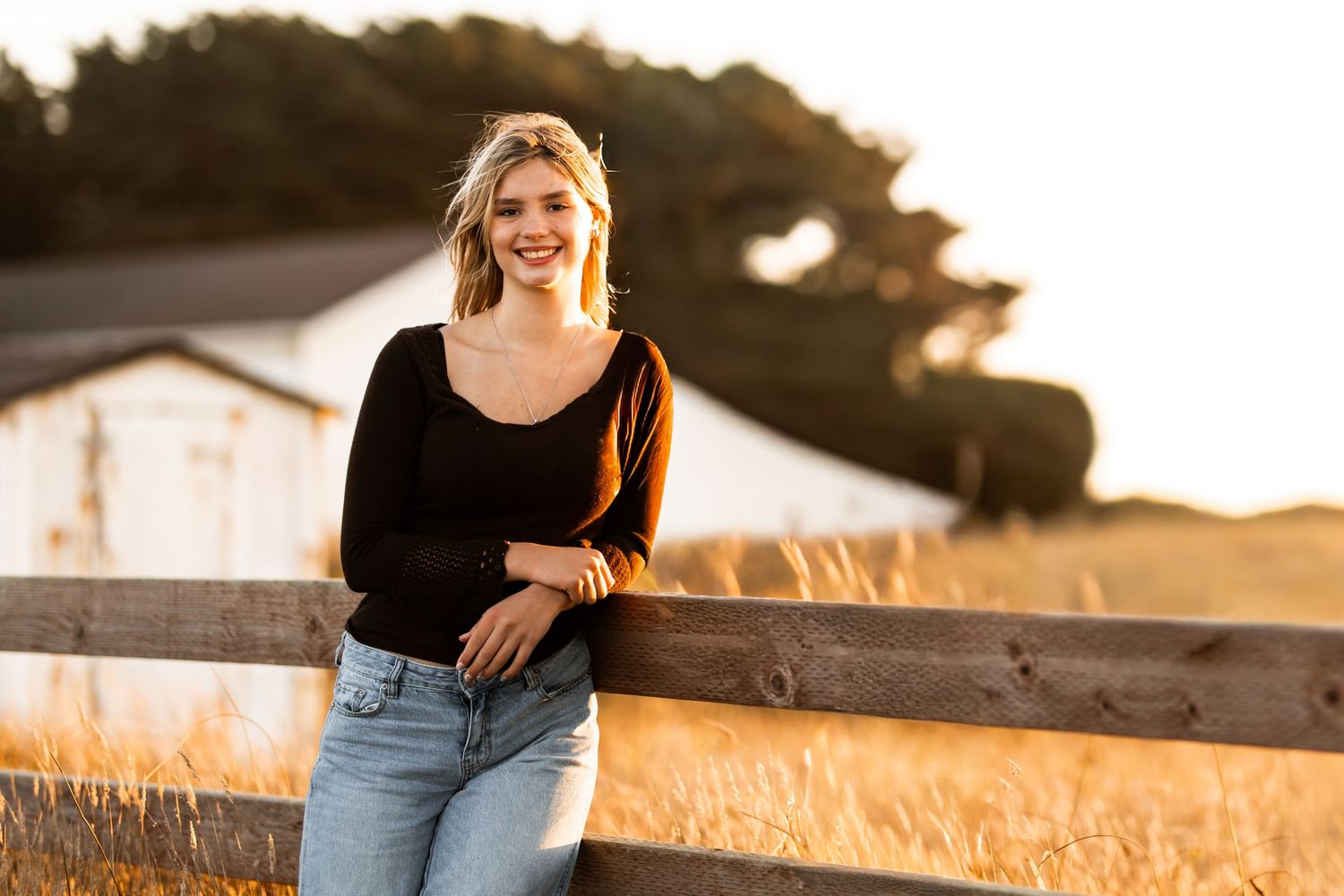 Smiling blonde teen girl leaning on wooden fence in golden field wearing black top and jeans.