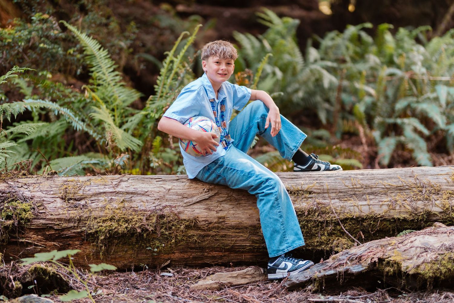 A boy sitting on a fallen log in a lush forest, wearing a blue shirt and jeans, holding a soccer ball.
