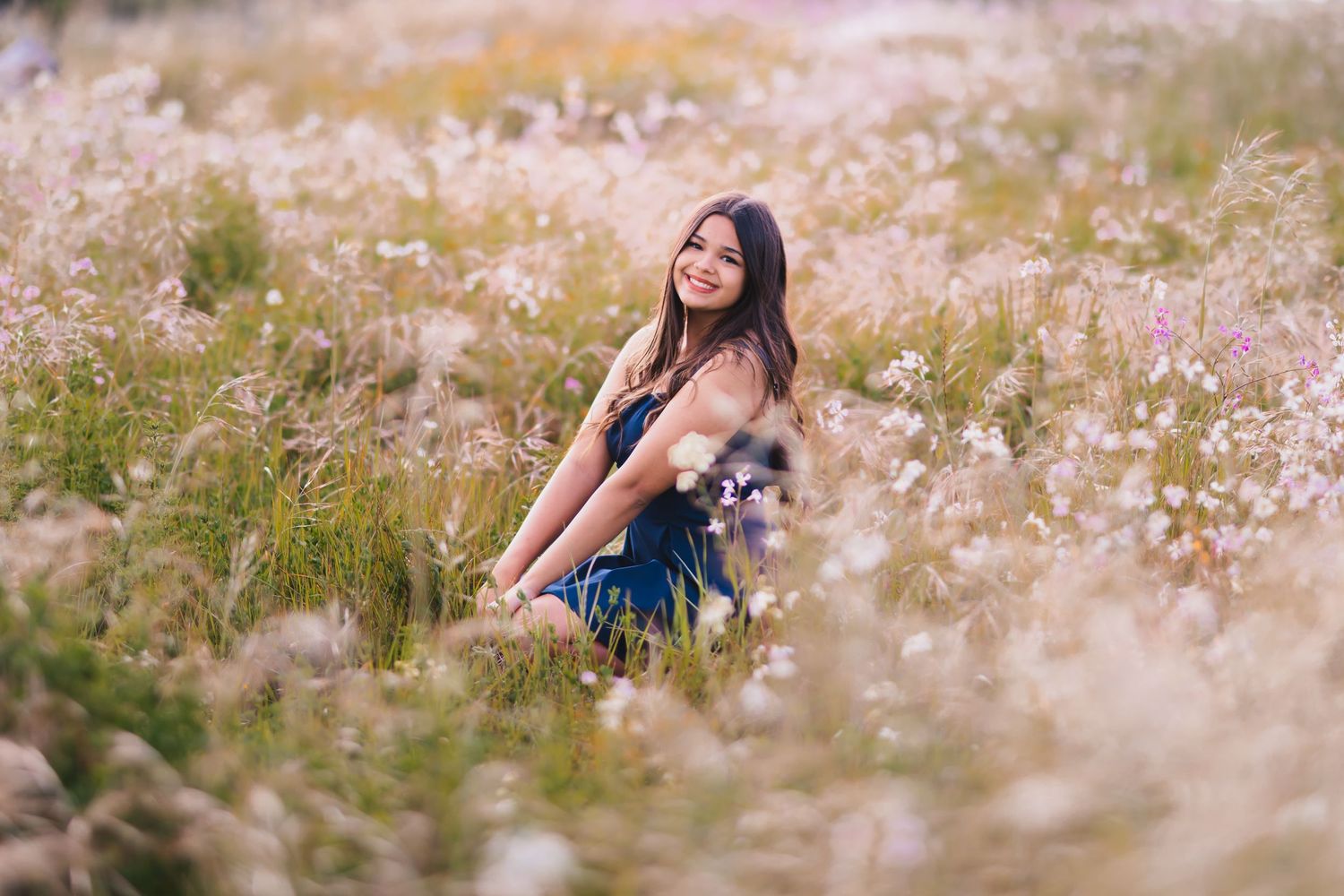 A smiling young woman in a blue dress sits among tall wildflowers in a dreamy, softly lit meadow.