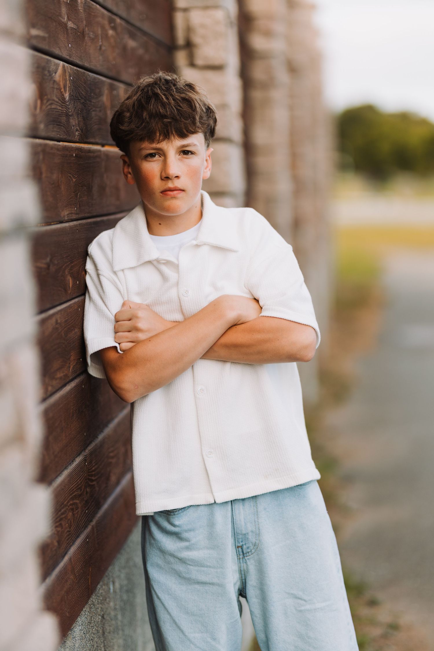 A teenage boy in a white shirt and jeans leans against a brick wall with arms crossed, looking serious outdoors.