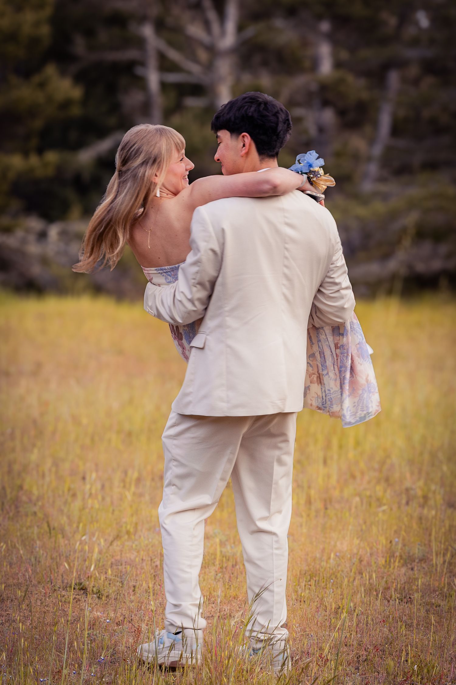 Prom couple embracing in golden field, girl in floral dress with arm around boy in tan suit.