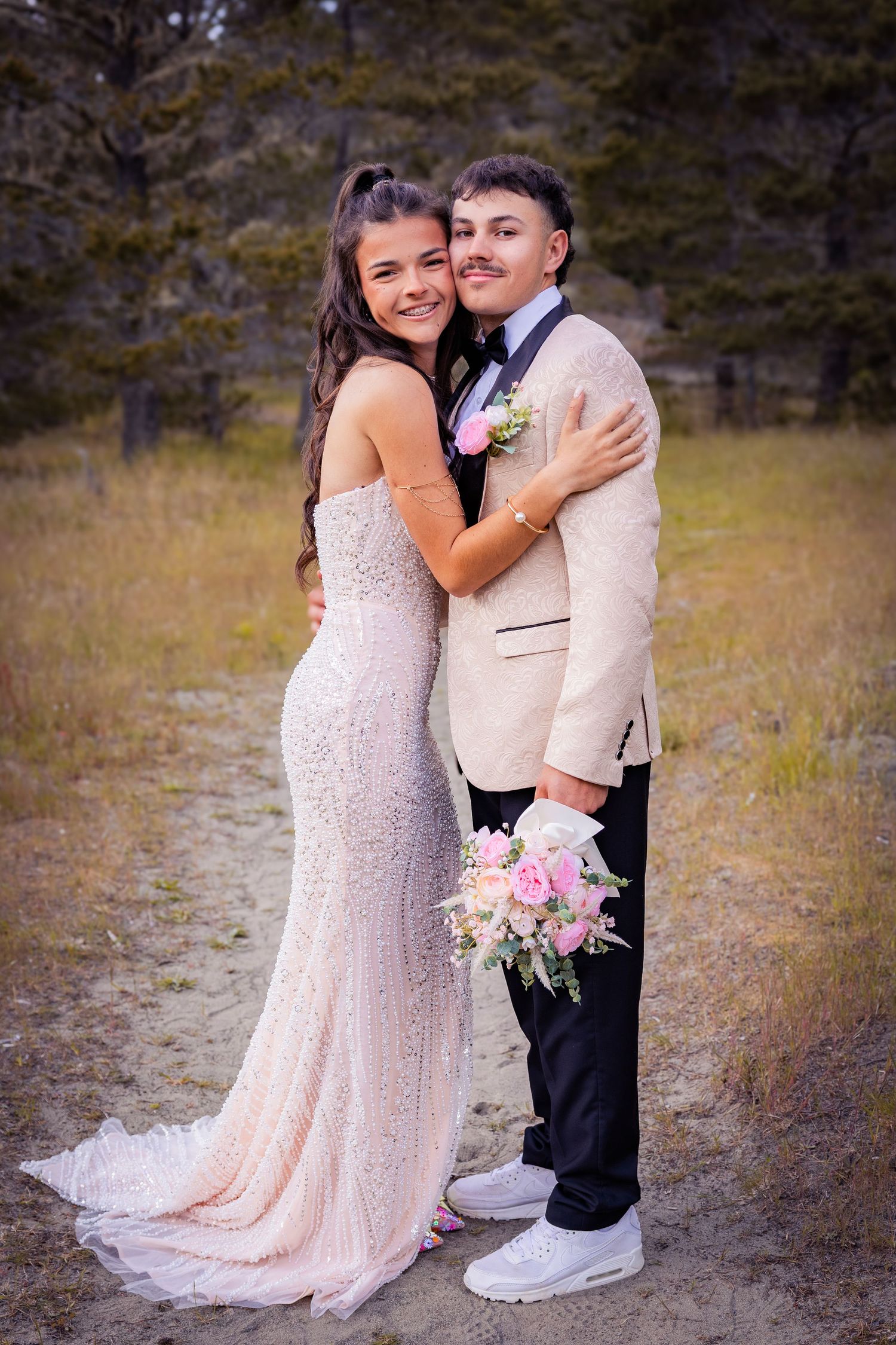 A young couple in formal prom attire poses outdoors, she in a sequined gown, he in a tan suit holding pink flowers.