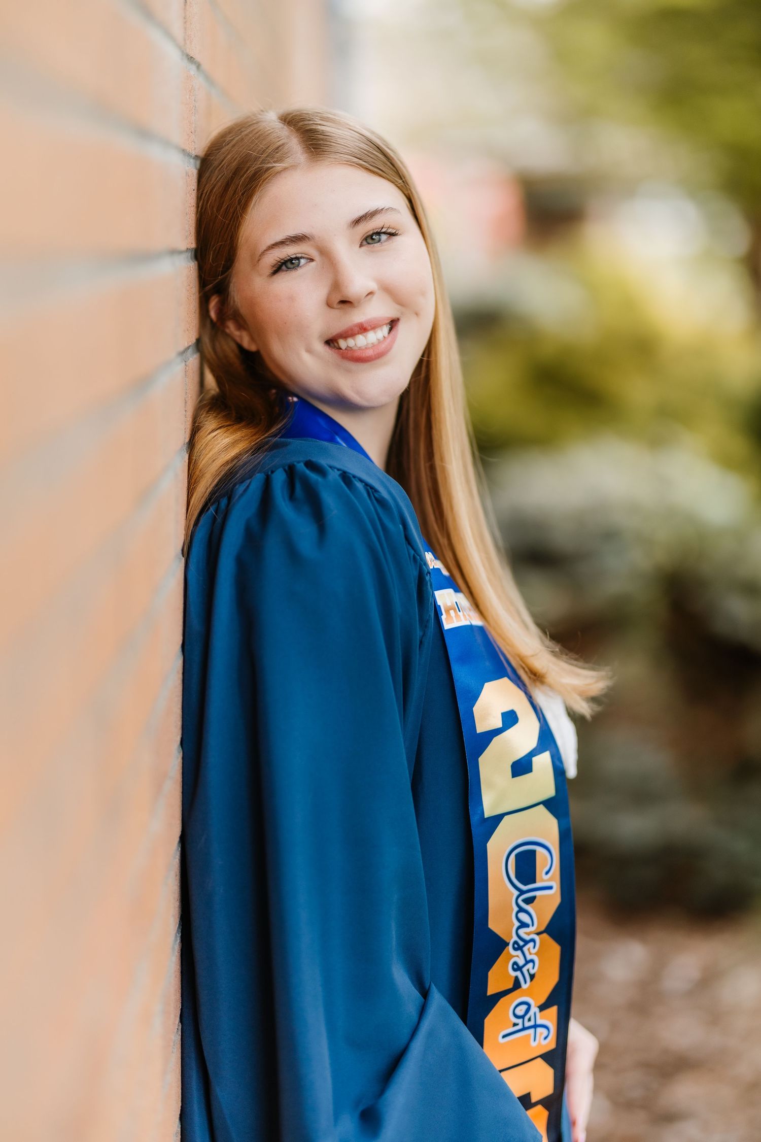 Smiling young woman in blue graduation gown and Class of sash leaning against brick wall outdoors.