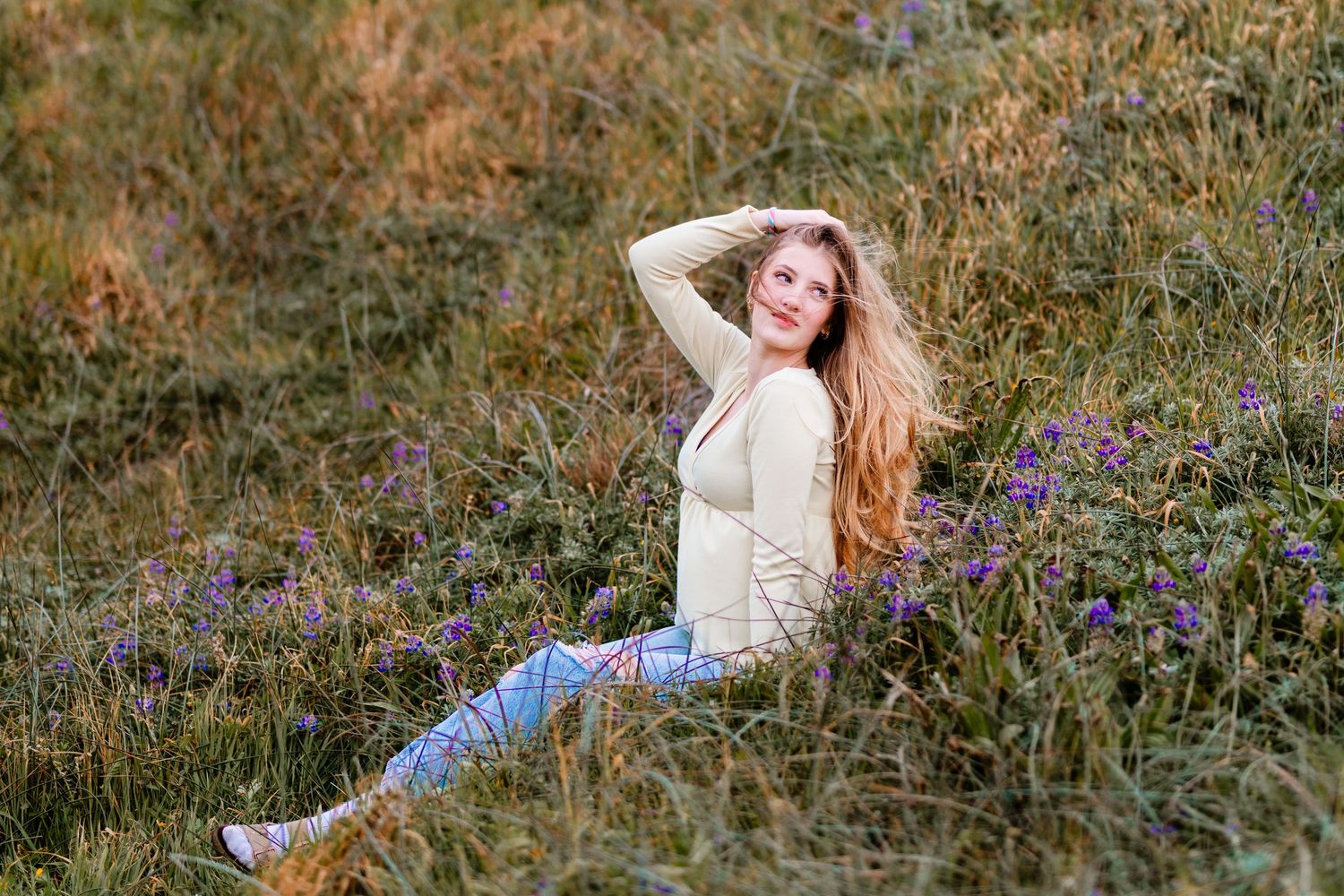 Young woman in cream cardigan and jeans sitting in a field of purple wildflowers.