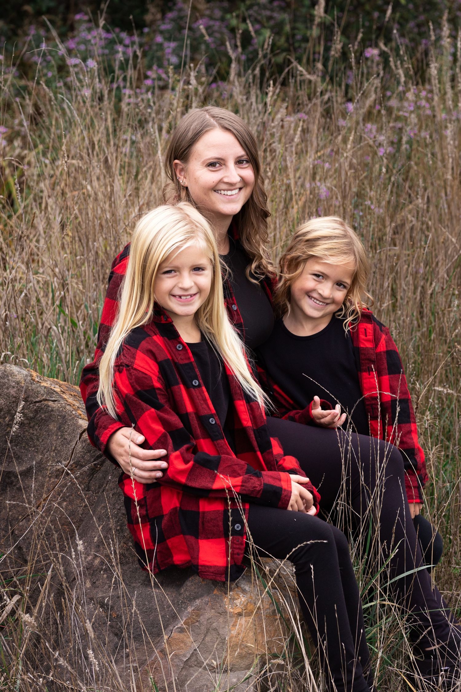 A smiling mother and two blonde daughters in matching red plaid shirts pose on rocks in a field.