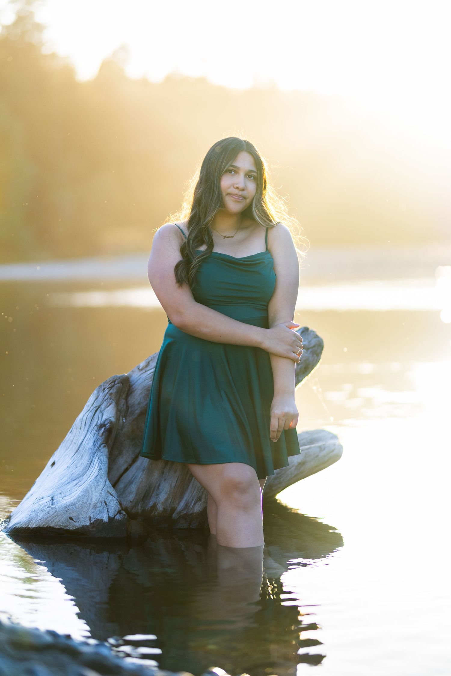 Young woman in teal dress standing in sunlit river beside driftwood log during golden hour.