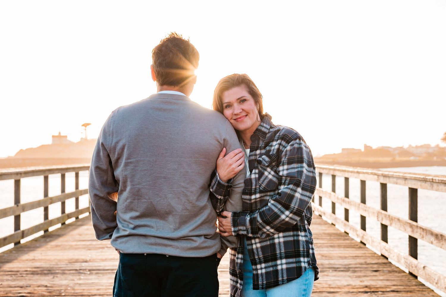 Woman in plaid shirt smiling while embracing a man from behind on a sunlit wooden pier at sunset.