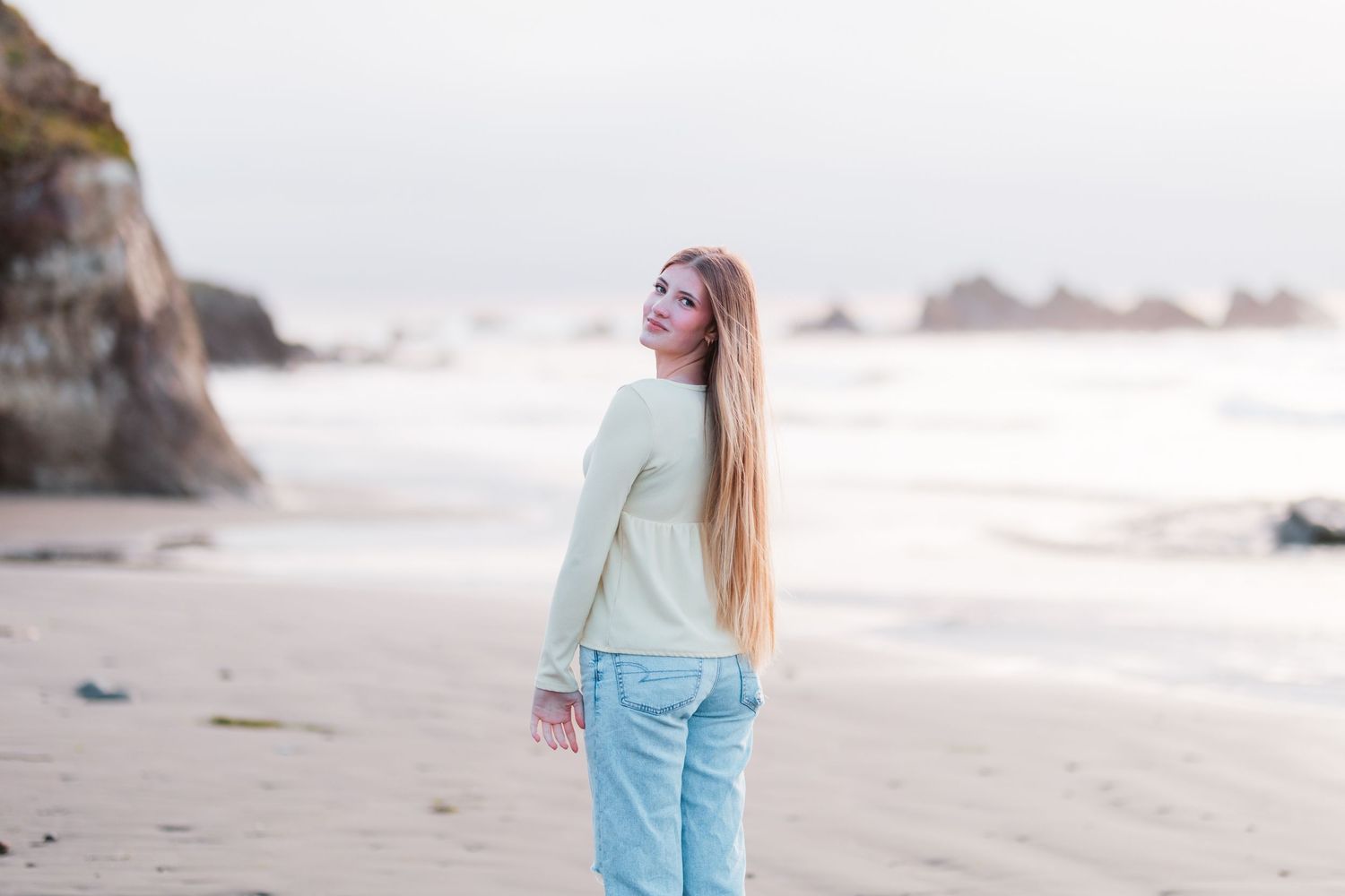 Young woman with long auburn hair in cream sweater walking on a scenic beach.