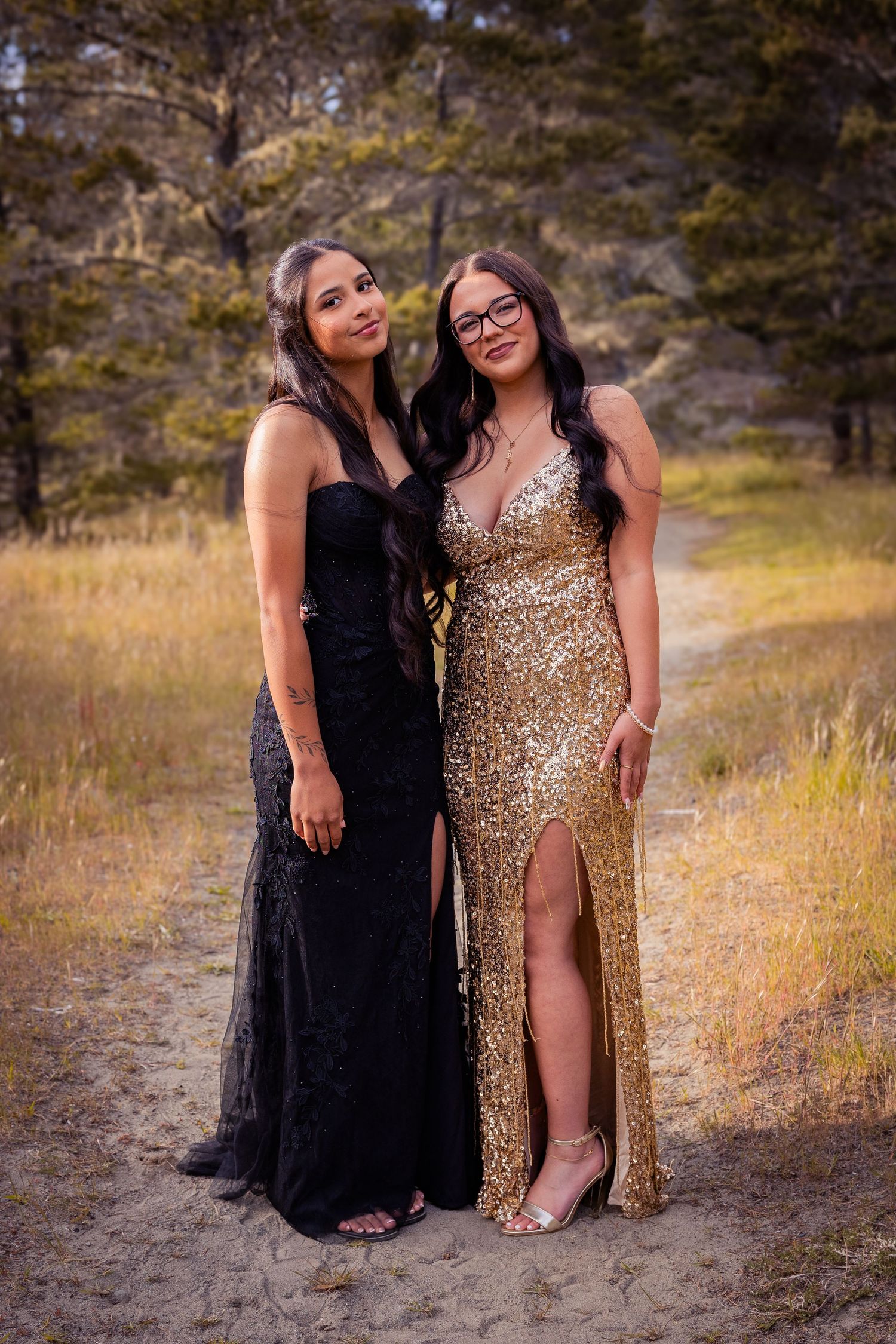 Two young women pose in formal gowns outdoors, one in black lace and one in a gold sequin dress with slit.